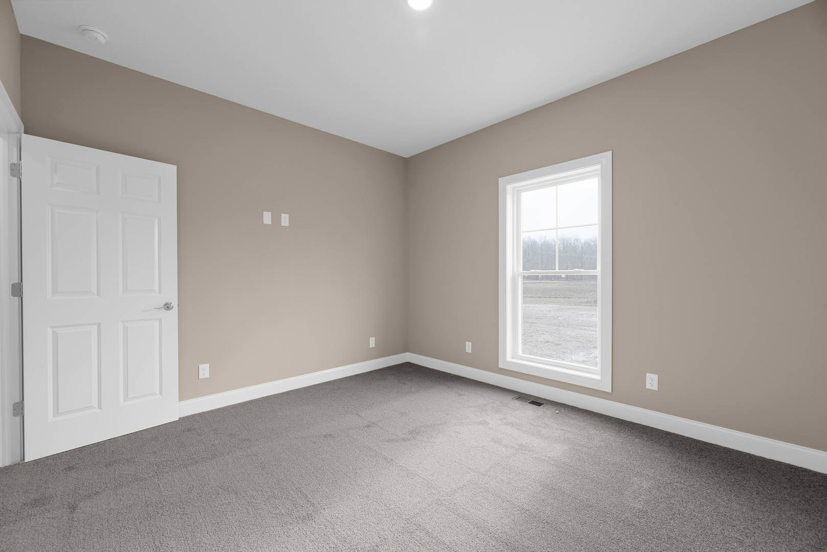 Bedroom with beige carpet, white walls, large window overlooking grassy field, white door with silver handle, crown molding, and ceiling with recessed lighting
