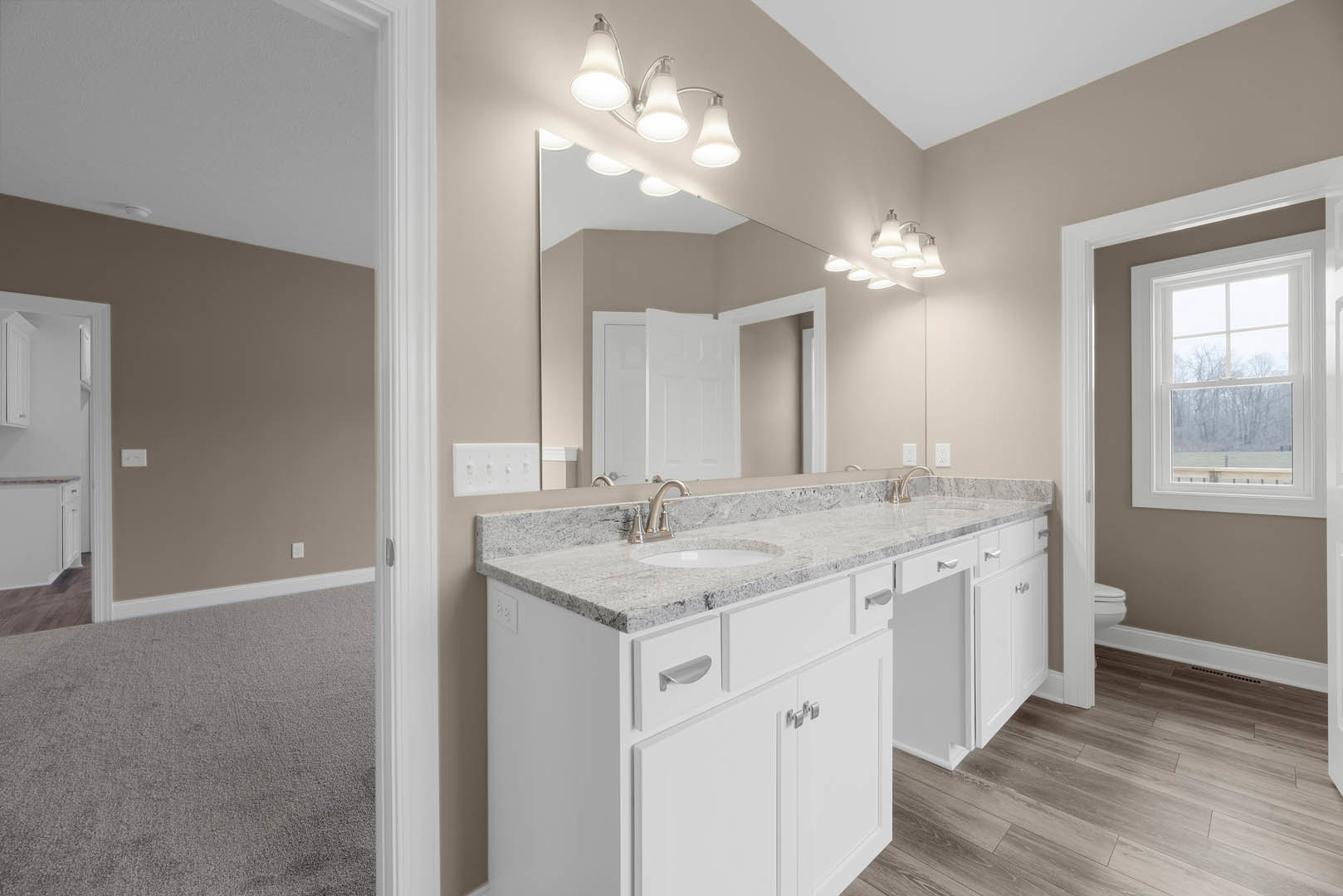 Bathroom featuring a wide marble countertop, large mirror above sink, white cabinetry, and window overlooking trees; three-light fixture mounted above mirror, white door, and tiled