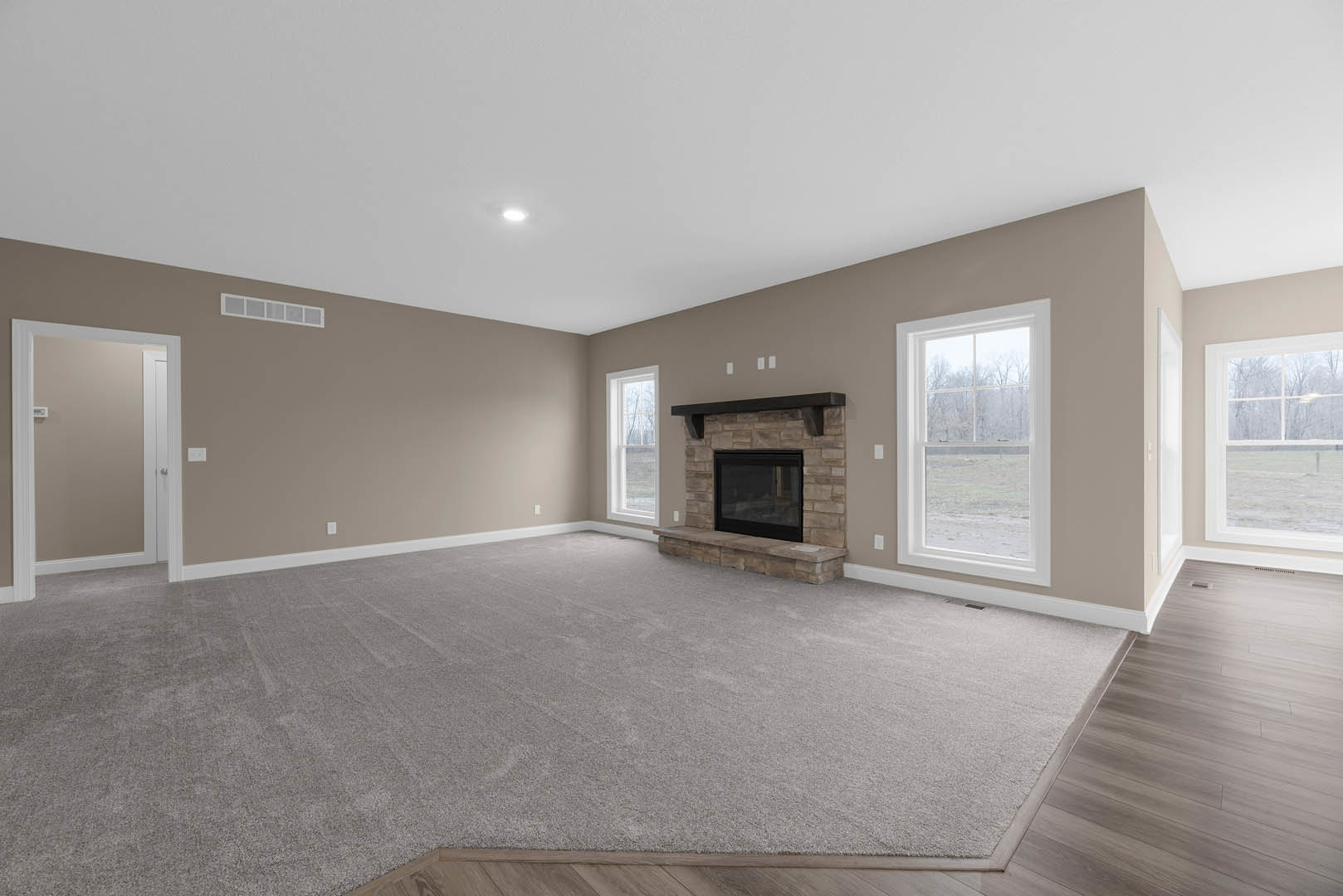 Living room with light carpet flooring, brick fireplace featuring a black glass door, large rectangular window overlooking a field, white door with matching frame, plaster ceiling