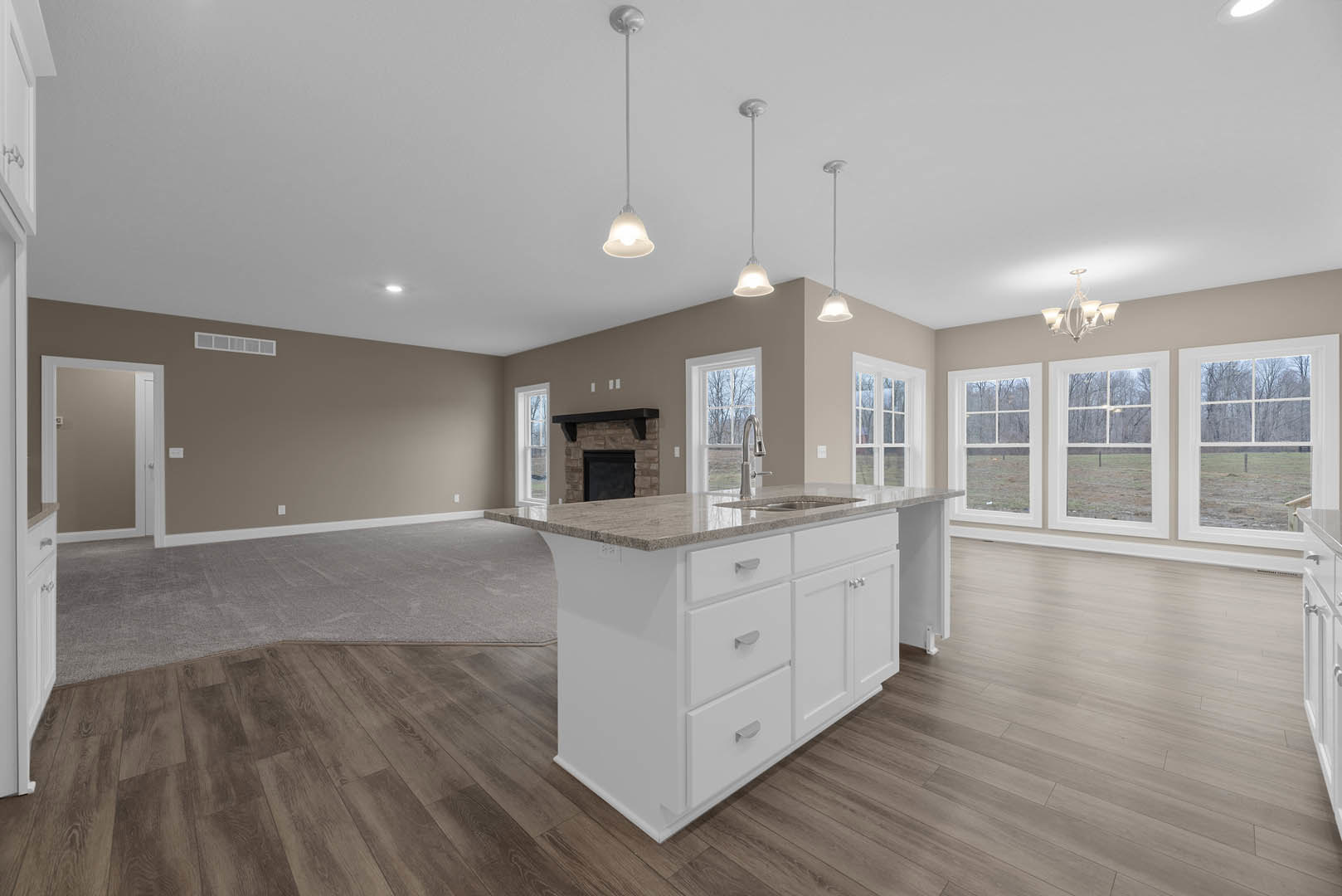 Open kitchen and living room featuring hardwood flooring, white kitchen island with built-in sink, white-framed door, row of windows overlooking trees, white walls with black trim