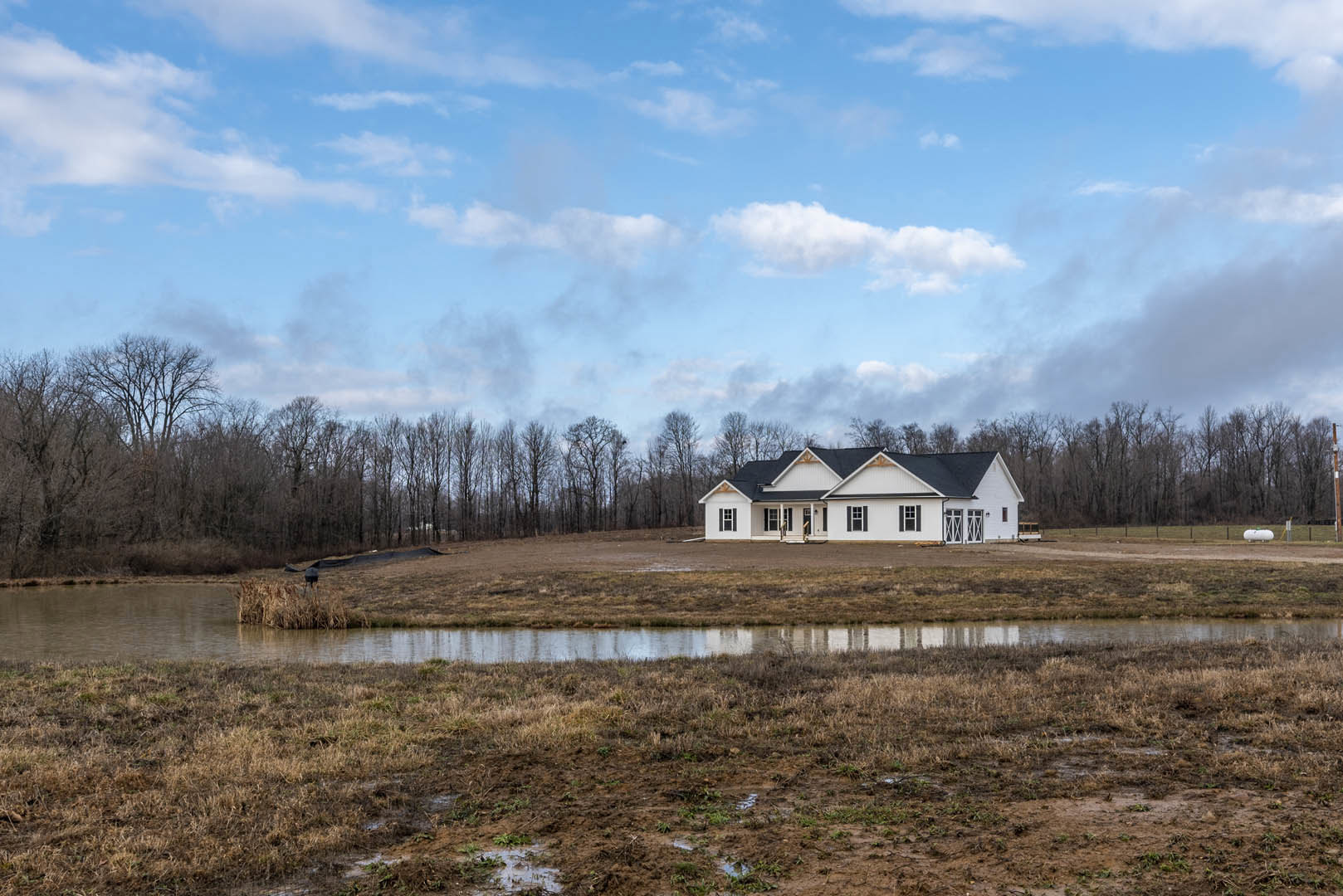 Modern house with black roof surrounded by grassy field, muddy patches, and scattered trees under cloudy sky