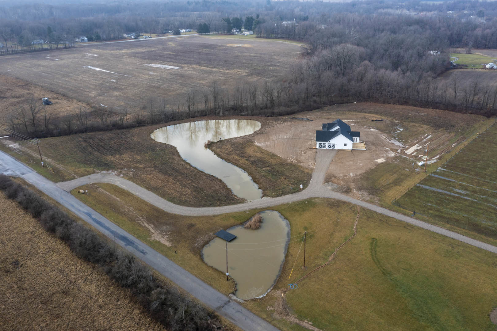 White house with black roof situated beside a small pond, surrounded by grassy plain and scattered trees, viewed from above under a clear sky