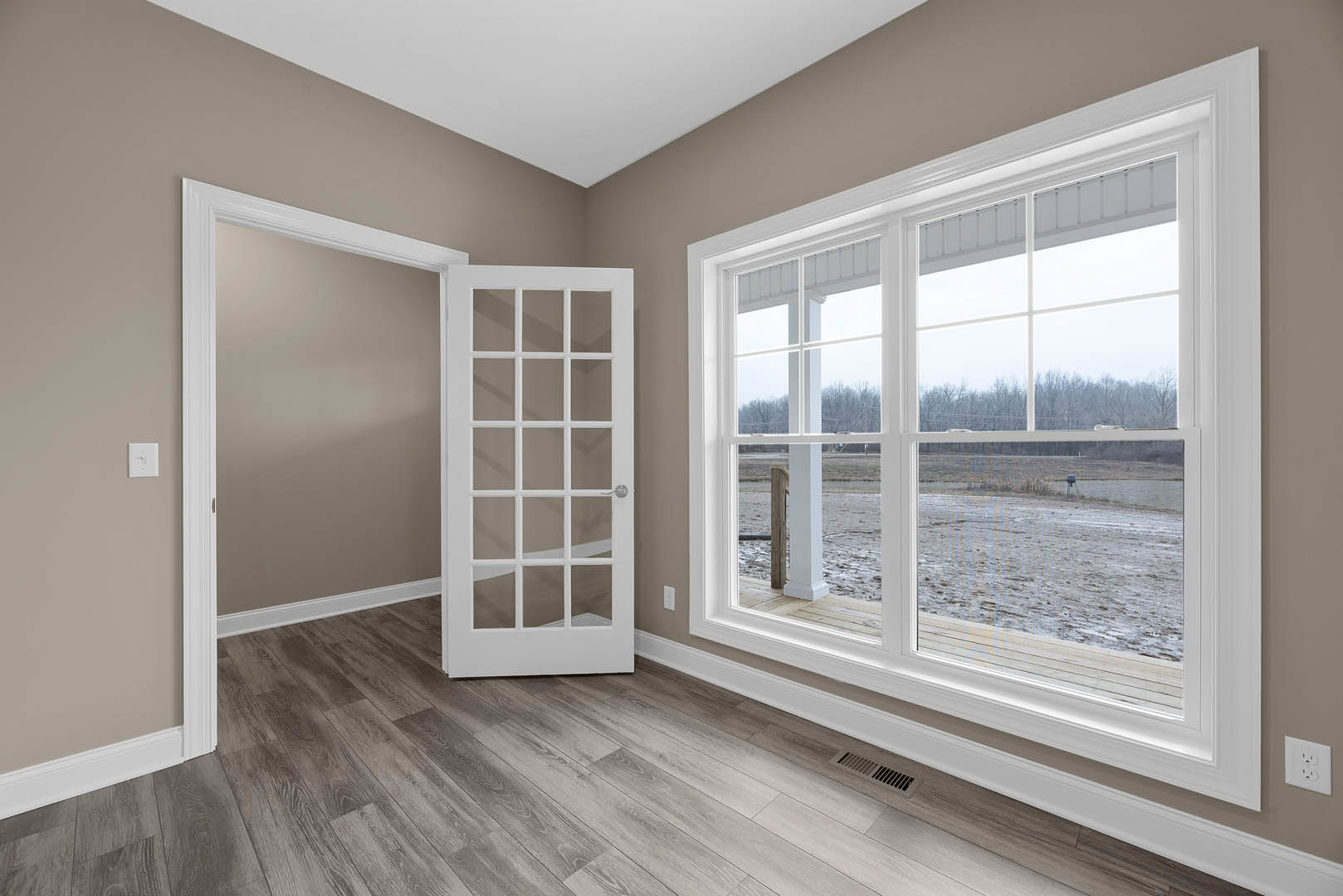 Wood floor with white door featuring glass panes, large window, white wall outlet, and floor vent in a bright interior room