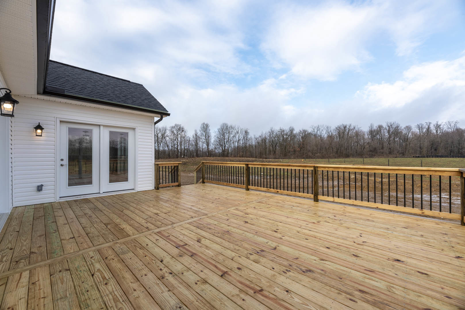 Wooden deck with black metal railings, white double doors, white-framed window, white siding, and leafy trees in the background