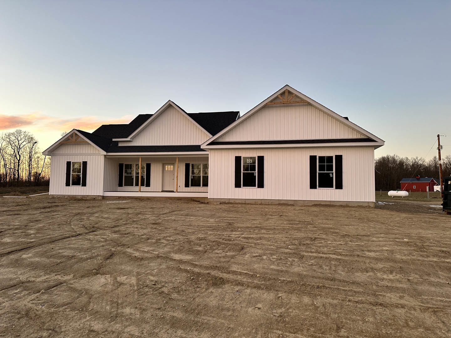 White house with black roof and black shutters, dirt yard with tire tracks, tree in background