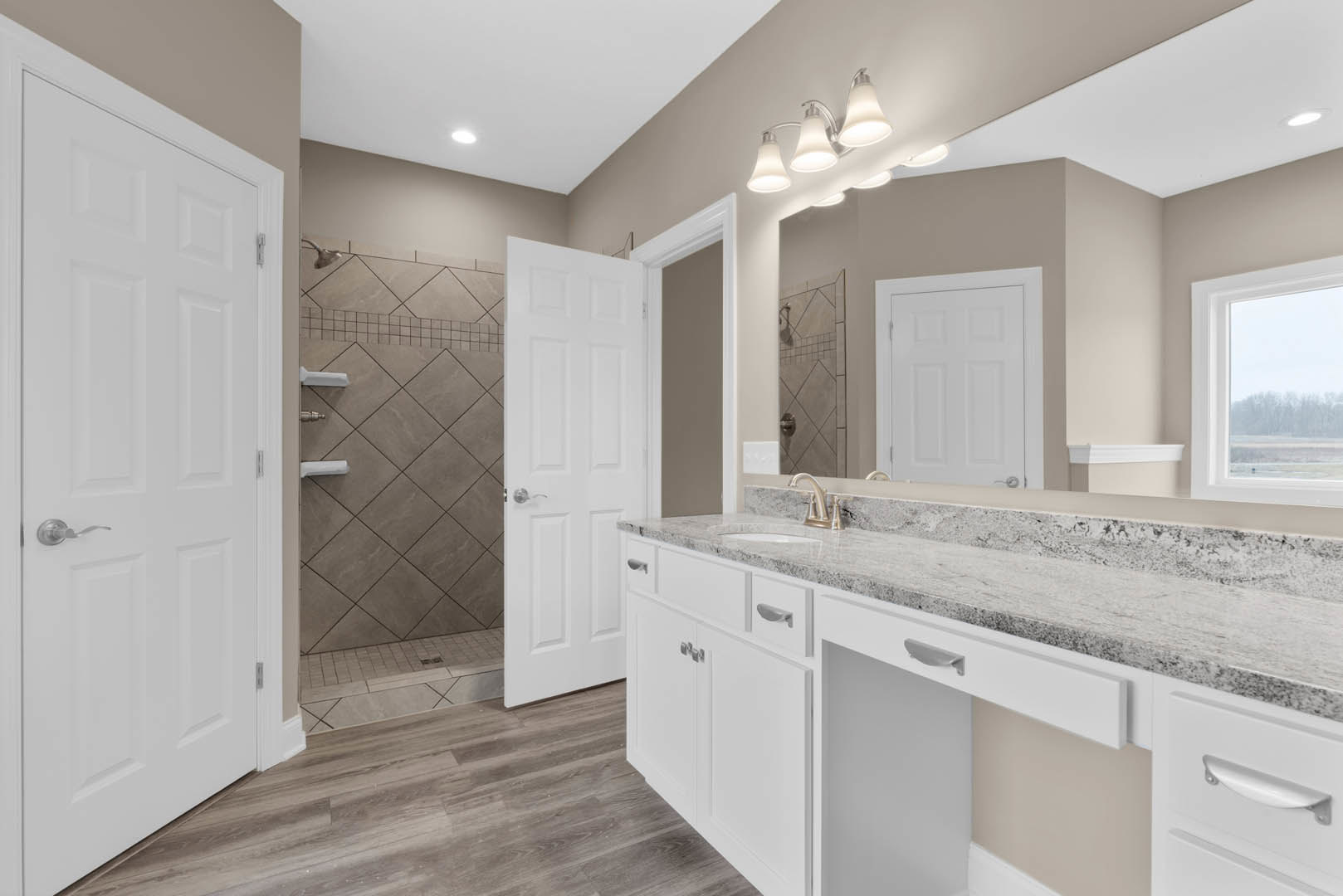 Bathroom featuring a marble countertop vanity, glass-enclosed shower with tile walls, white door with silver handle, window overlooking trees, and modern light fixture.