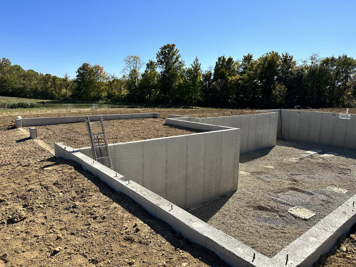 Concrete foundation with a metal ladder positioned at the center, surrounded by dirt and bordered by trees under a clear blue sky