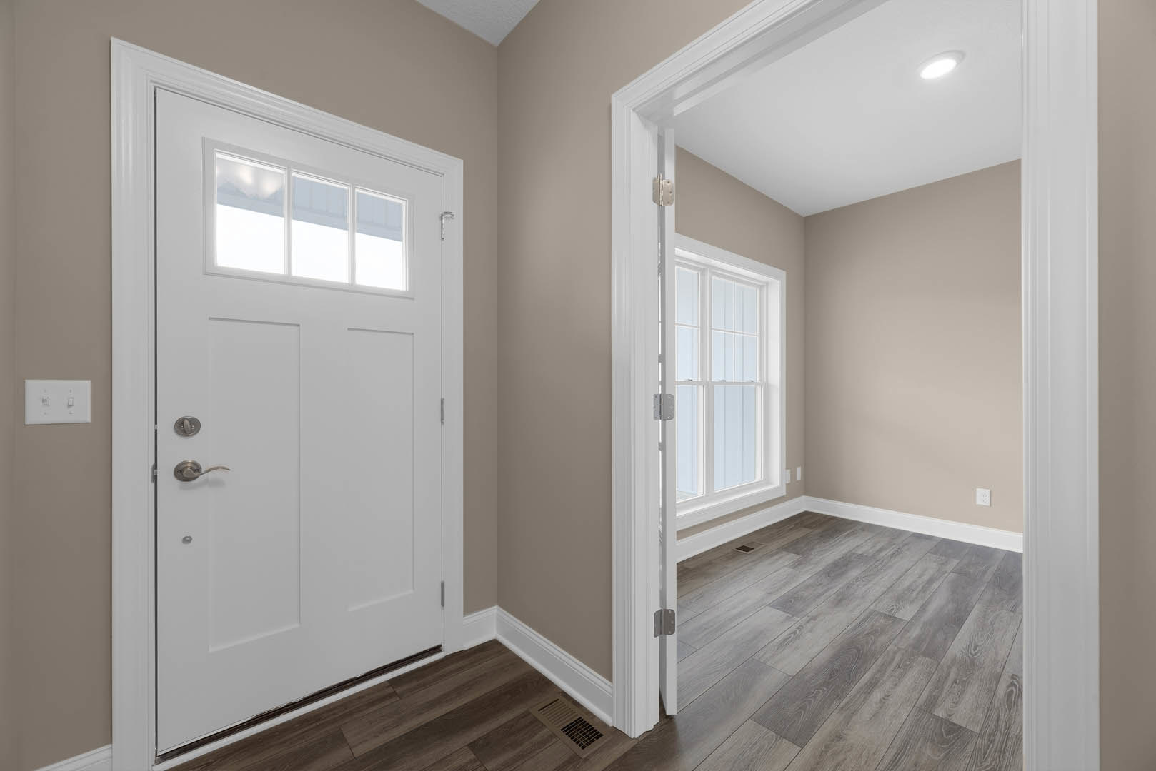 Hallway with white paneled door, brushed metal handle, rectangular window, laminate wood flooring, white plaster walls, ceiling light fixture, and double light switch.