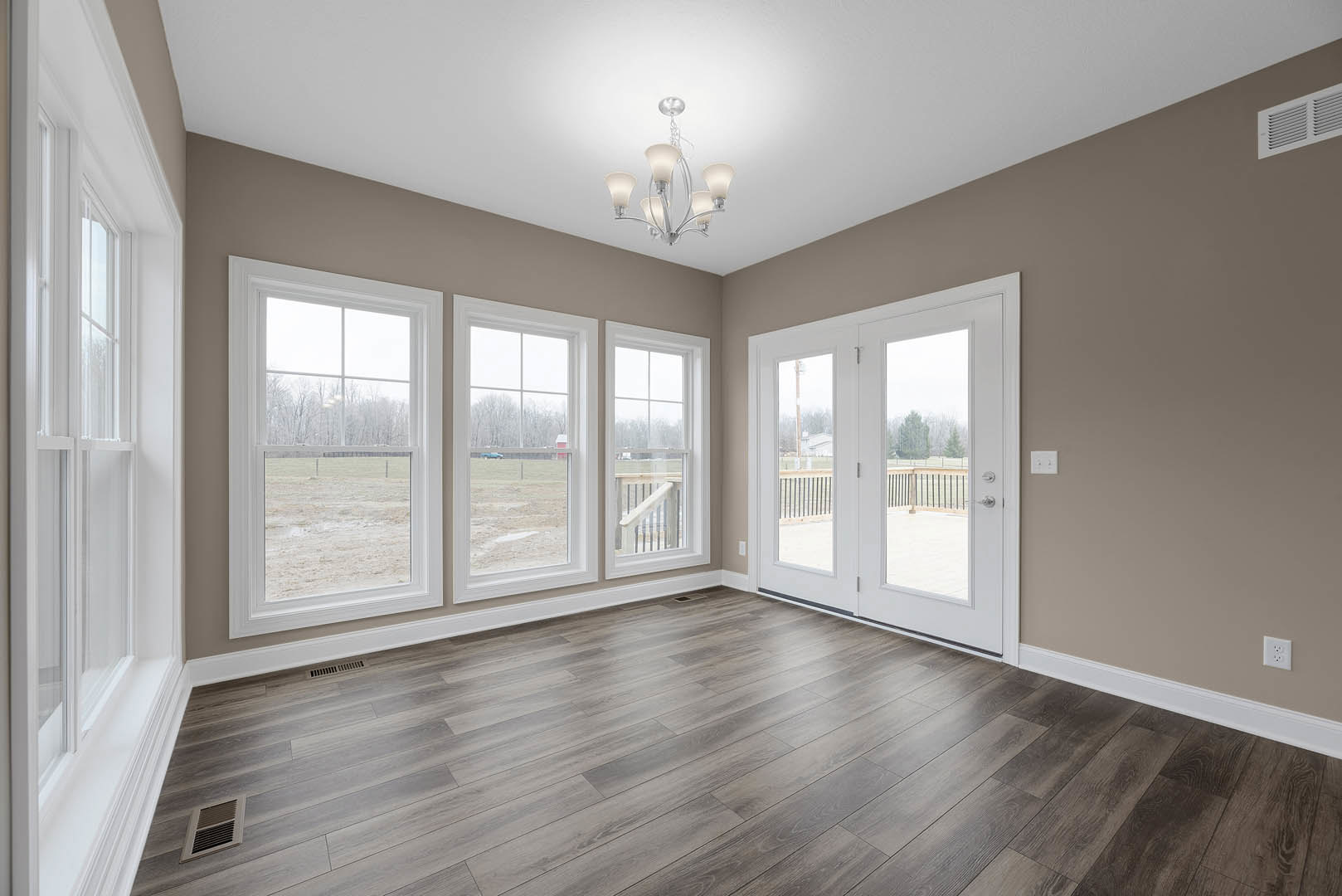 Wood flooring in a spacious room with a decorative chandelier, white double doors with glass panes, large window overlooking a field, ceiling vent, and detailed wall molding