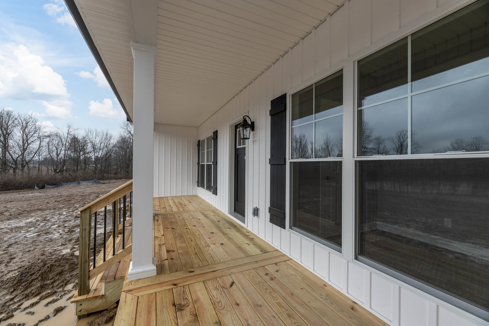 White siding house with large wooden deck, front porch, multiple windows, outdoor lamp, pitched roof, surrounded by trees under blue sky.