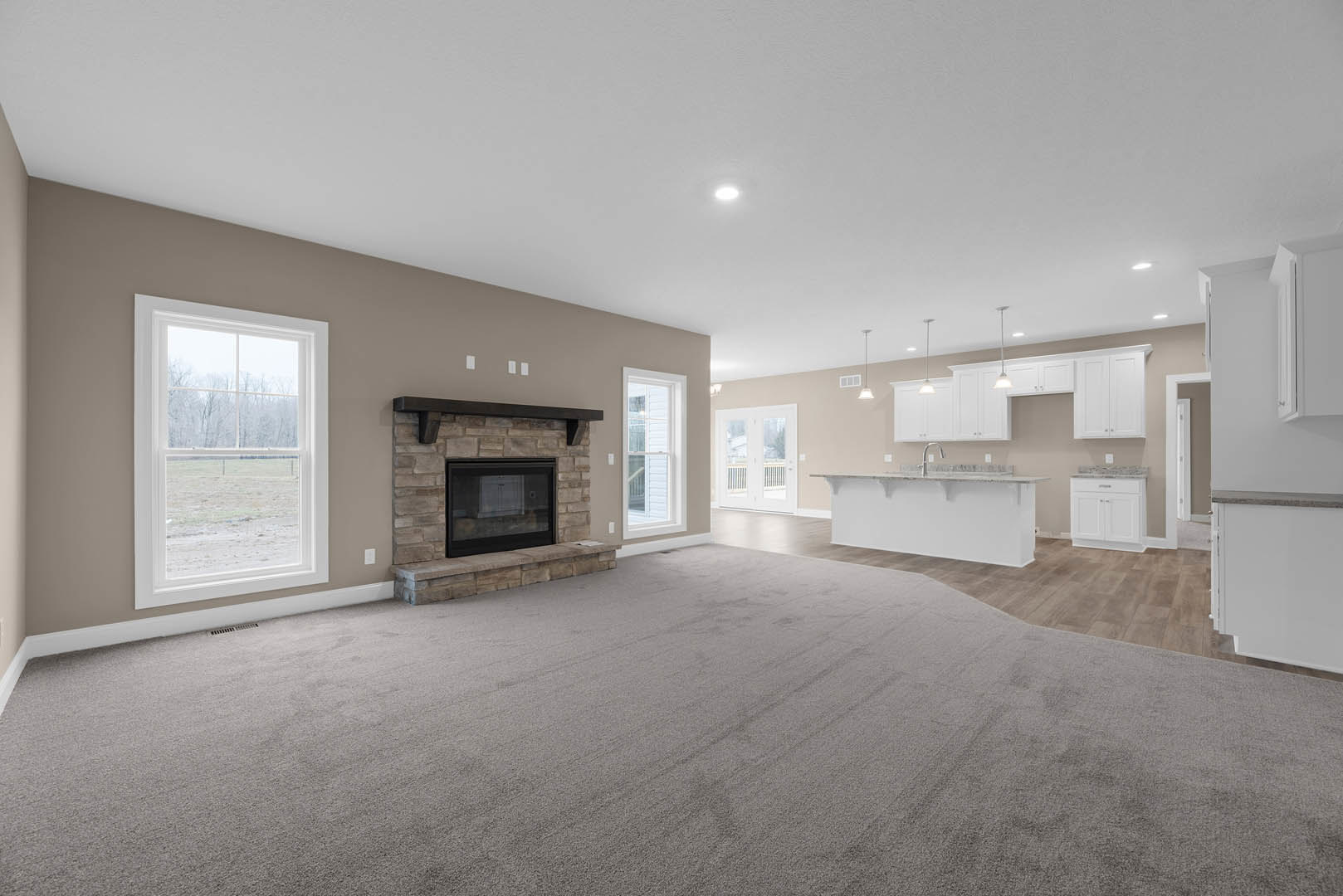 Open-concept living room with light wood flooring, white-framed windows overlooking a field, black glass fireplace surrounded by plaster walls, neutral carpet, and modern kitchen