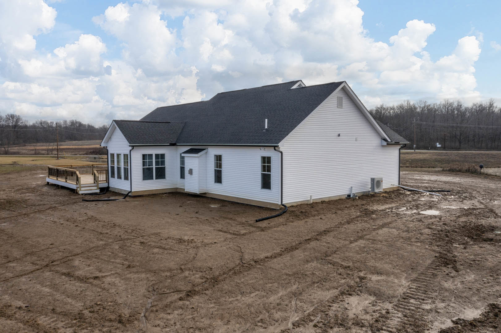 White house with black roof, wooden deck with metal railing, dirt yard with tire tracks, cloudy sky overhead