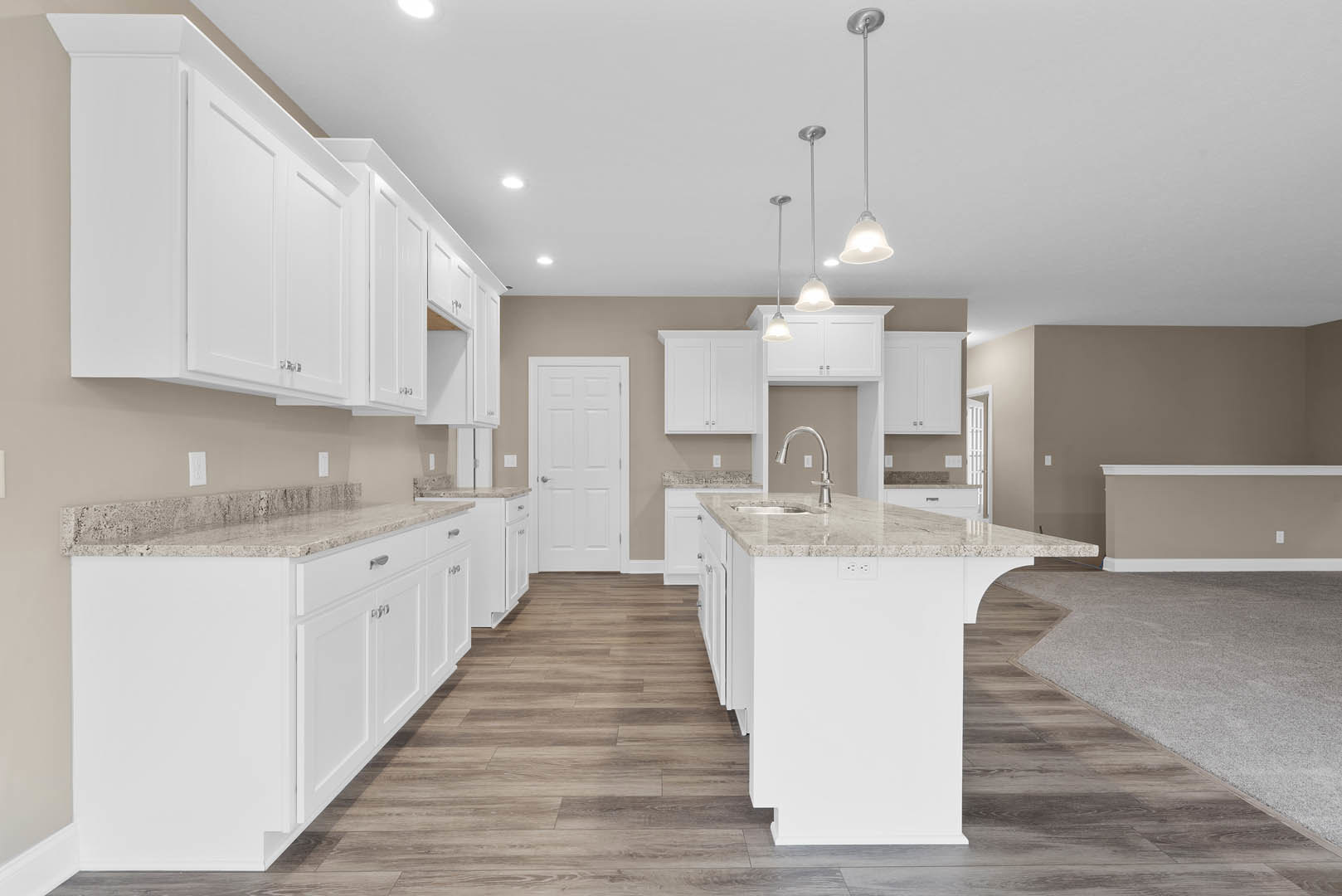 Kitchen with white shaker cabinets, wood plank flooring, white door with silver handle, marble-topped island, stainless steel sink, pendant lights, and neutral carpet visible in