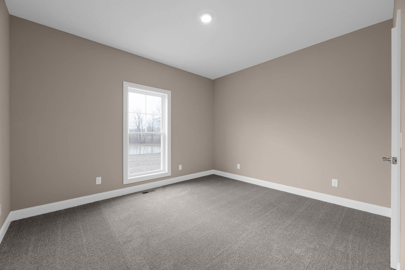 Bright room with white walls, grey carpet flooring, large window overlooking trees and pond, recessed ceiling light, simple molding.