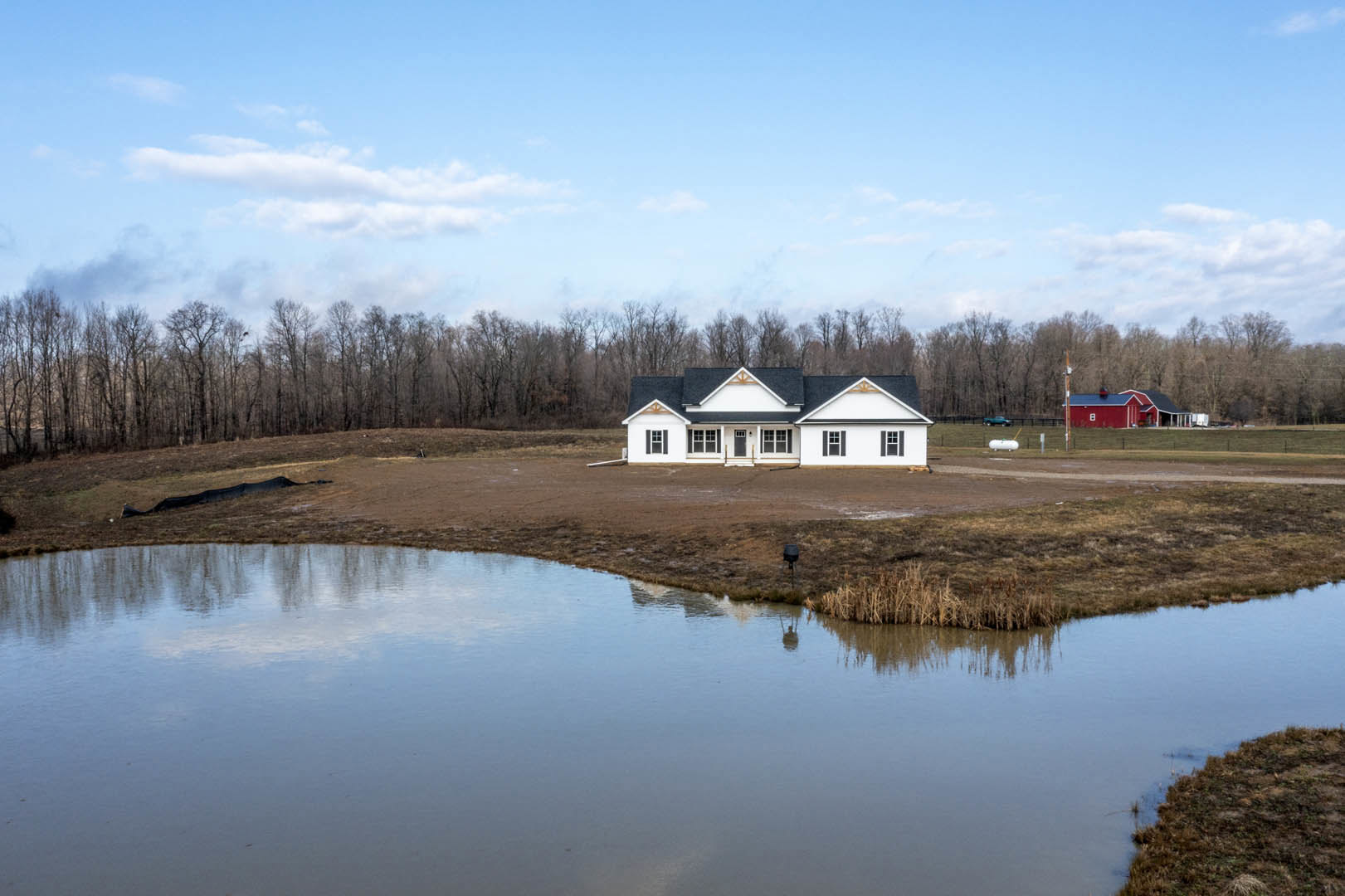 White house with black roof beside a lake, grassy shoreline, red barn with blue roof in background, trees under partly cloudy blue sky