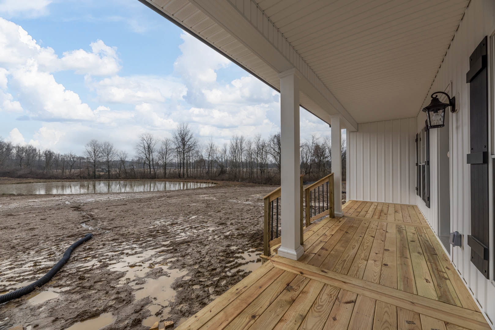 Covered porch with wooden deck, white pillars, and railings overlooking muddy ground with scattered black pipe and leafless trees in the background