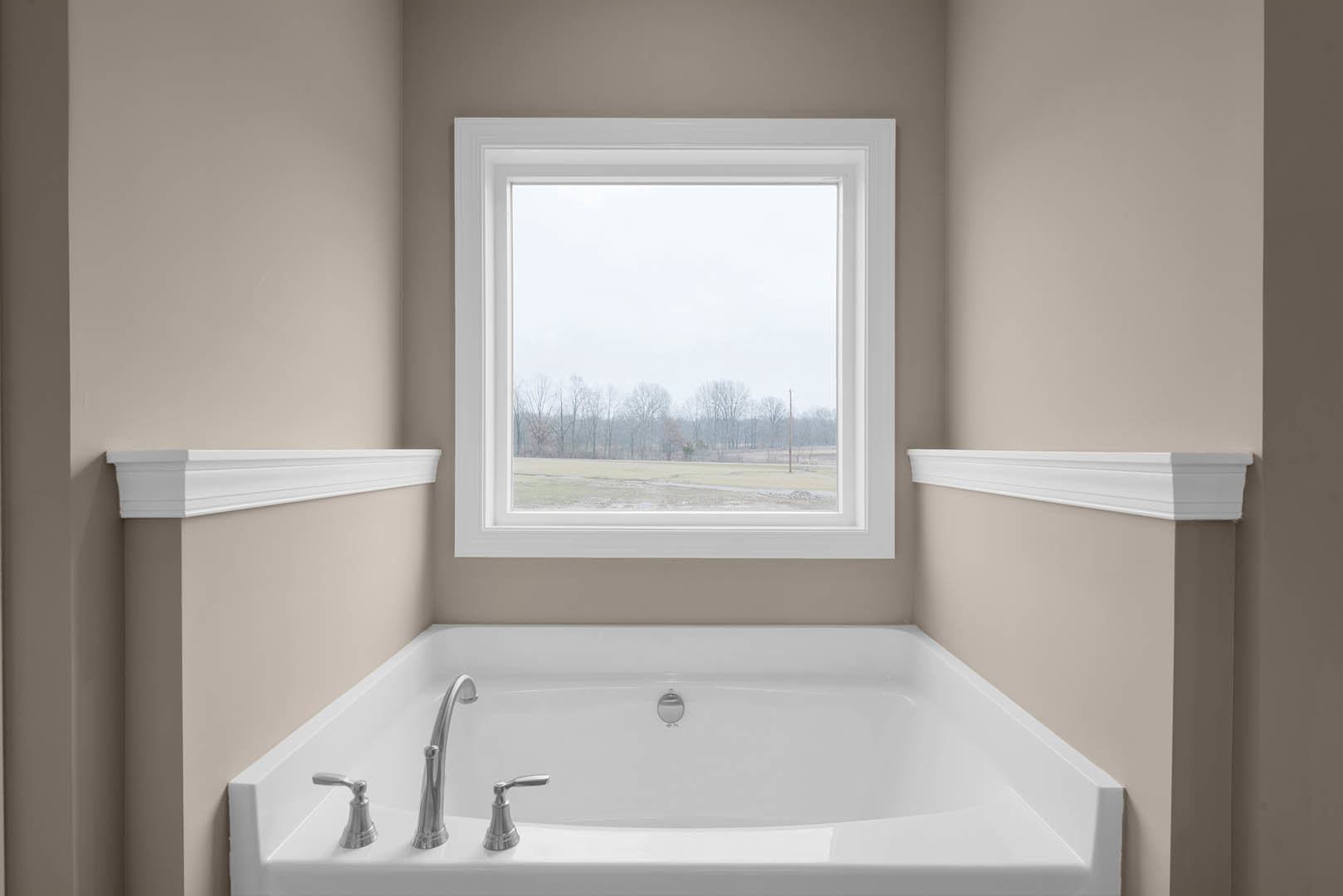 Freestanding white bathtub beneath large window overlooking foggy field and trees, chrome faucet, white wall molding