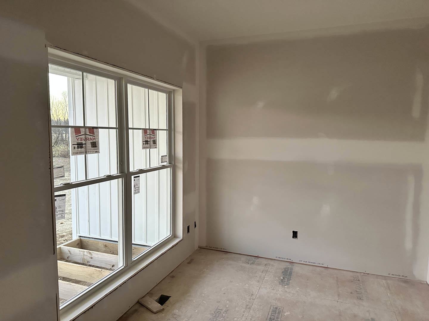 White plaster wall and ceiling, large window with wooden planks and daylight, light-colored floor, small signs visible on window glass
