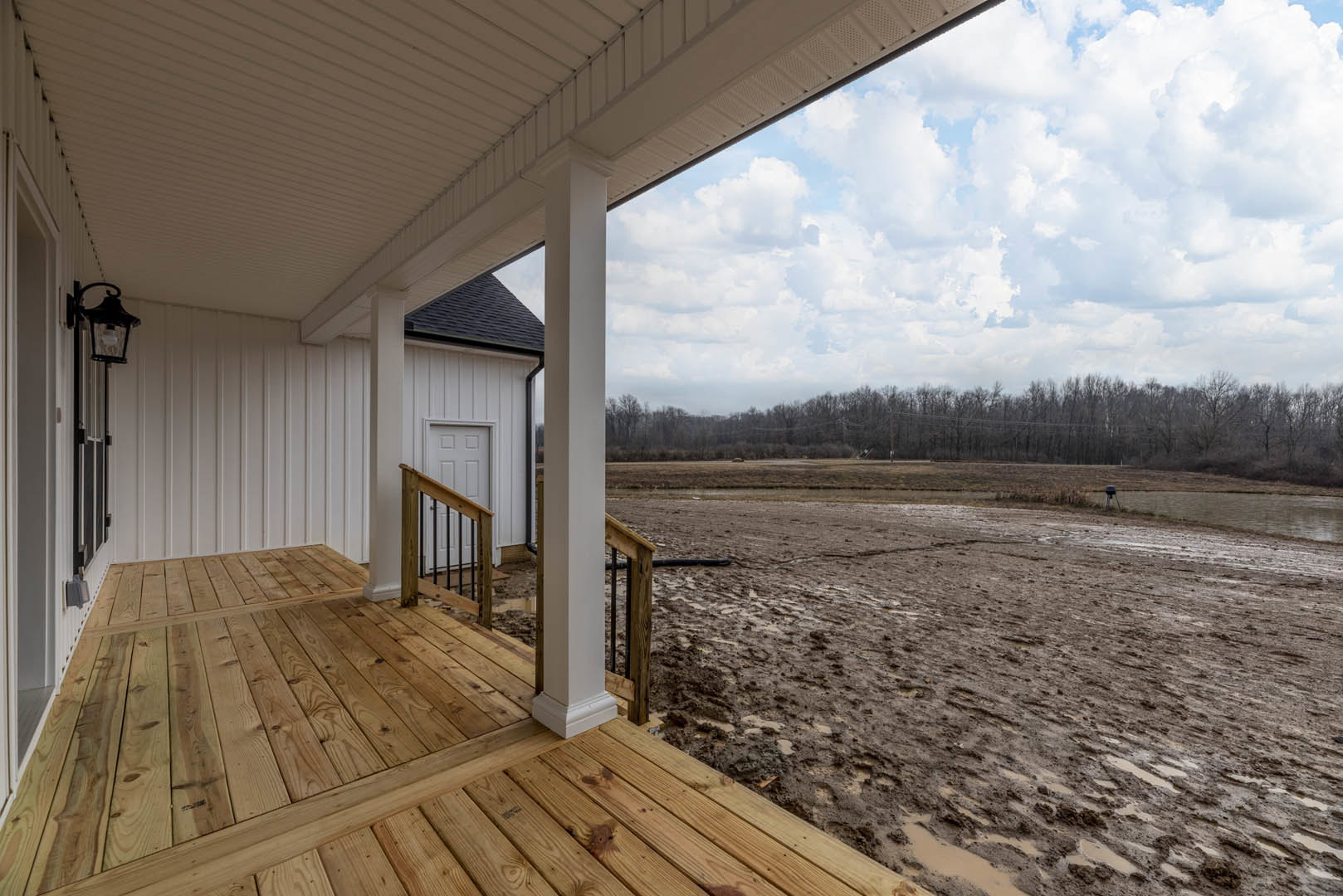 White-roofed house with wooden deck and metal gate, muddy field bordered by fence and distant tower, white pillar and wooden railing with metal bars, blue sky and scattered clouds