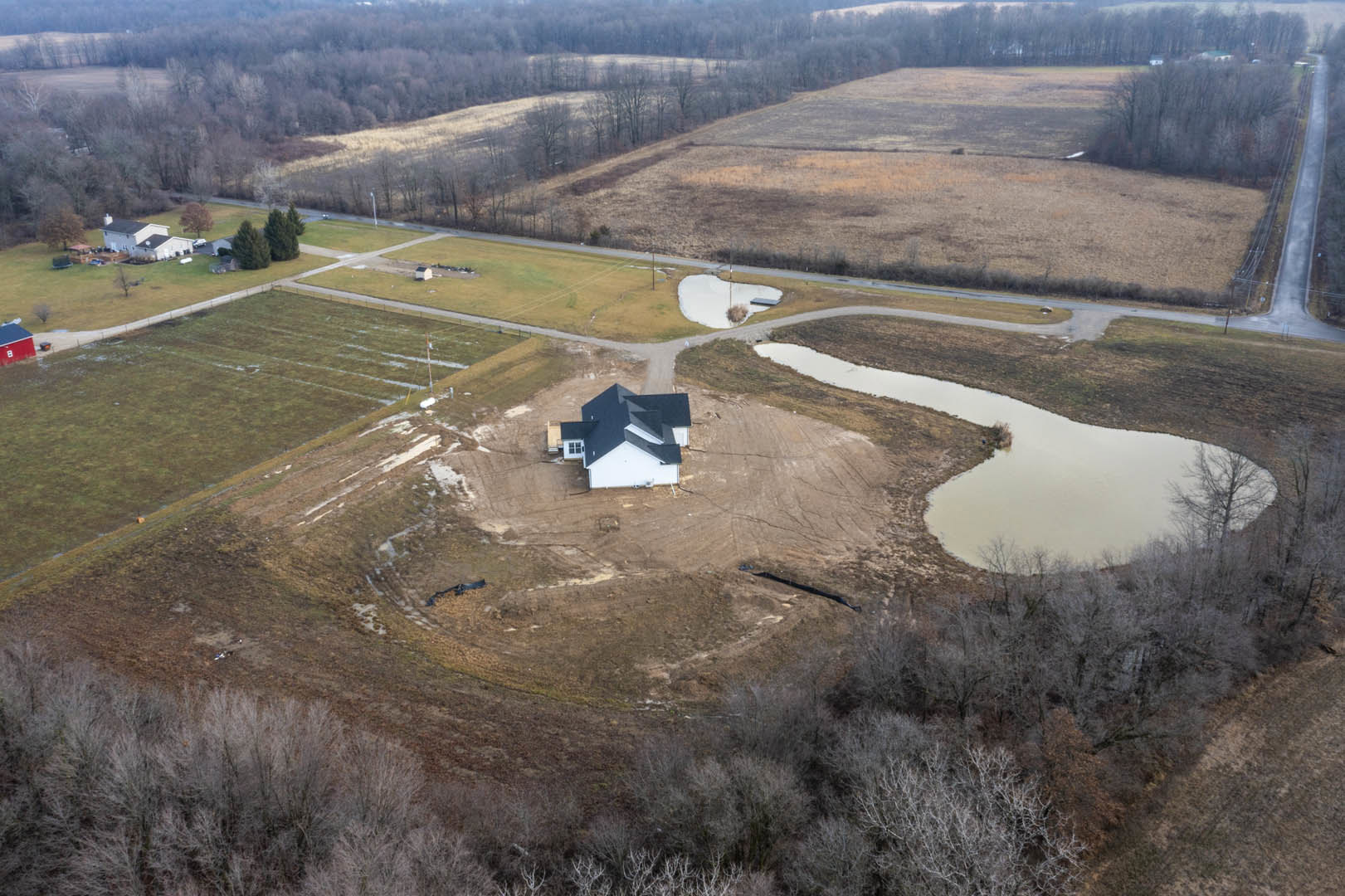 White house with black roof situated in a grassy field, small pond and tree visible in the background, mountain range on the horizon under a clear sky.
