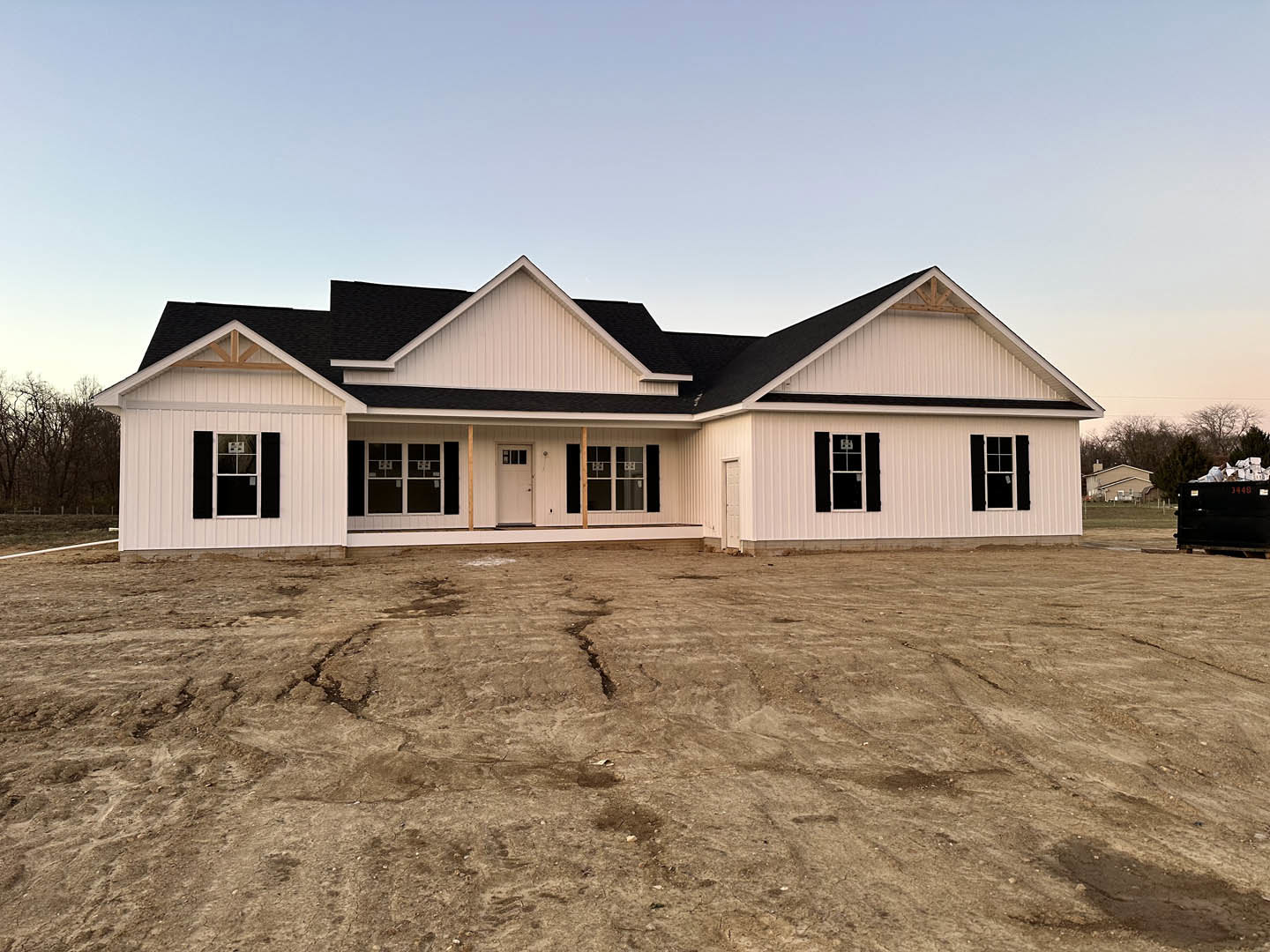 White house with black roof, white framed door, windows, dirt yard, large ground crack, black dumpster filled with boxes, surrounding trees.