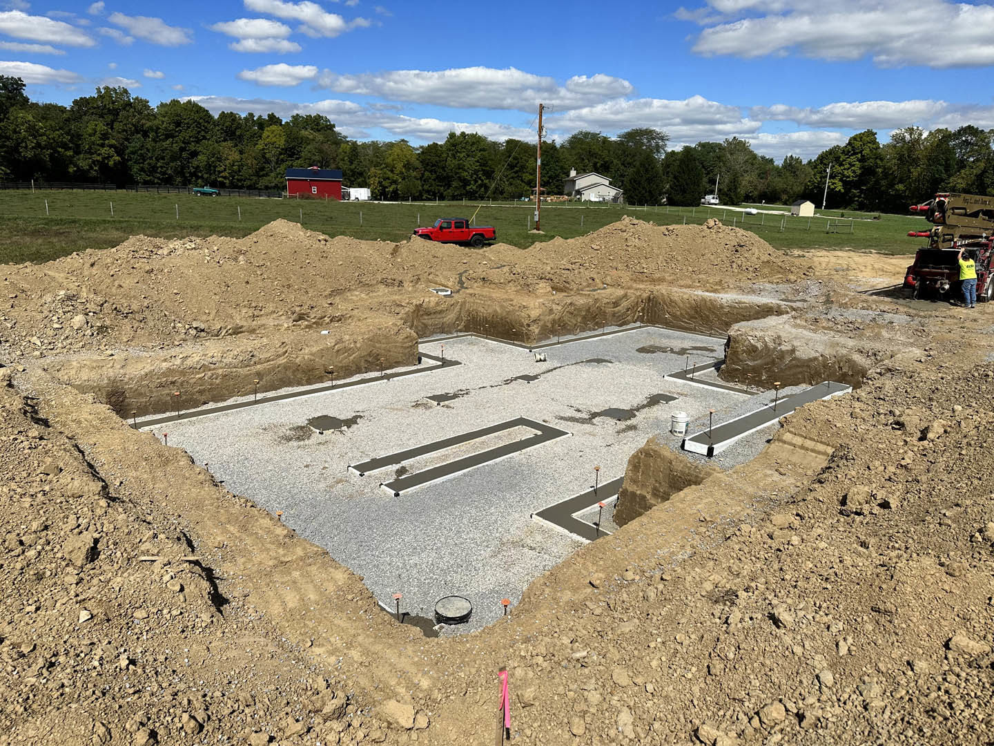 Construction site with exposed dirt, round tank being worked on by a man, red truck parked in background, red barn with blue roof, blue sky and scattered clouds overhead.