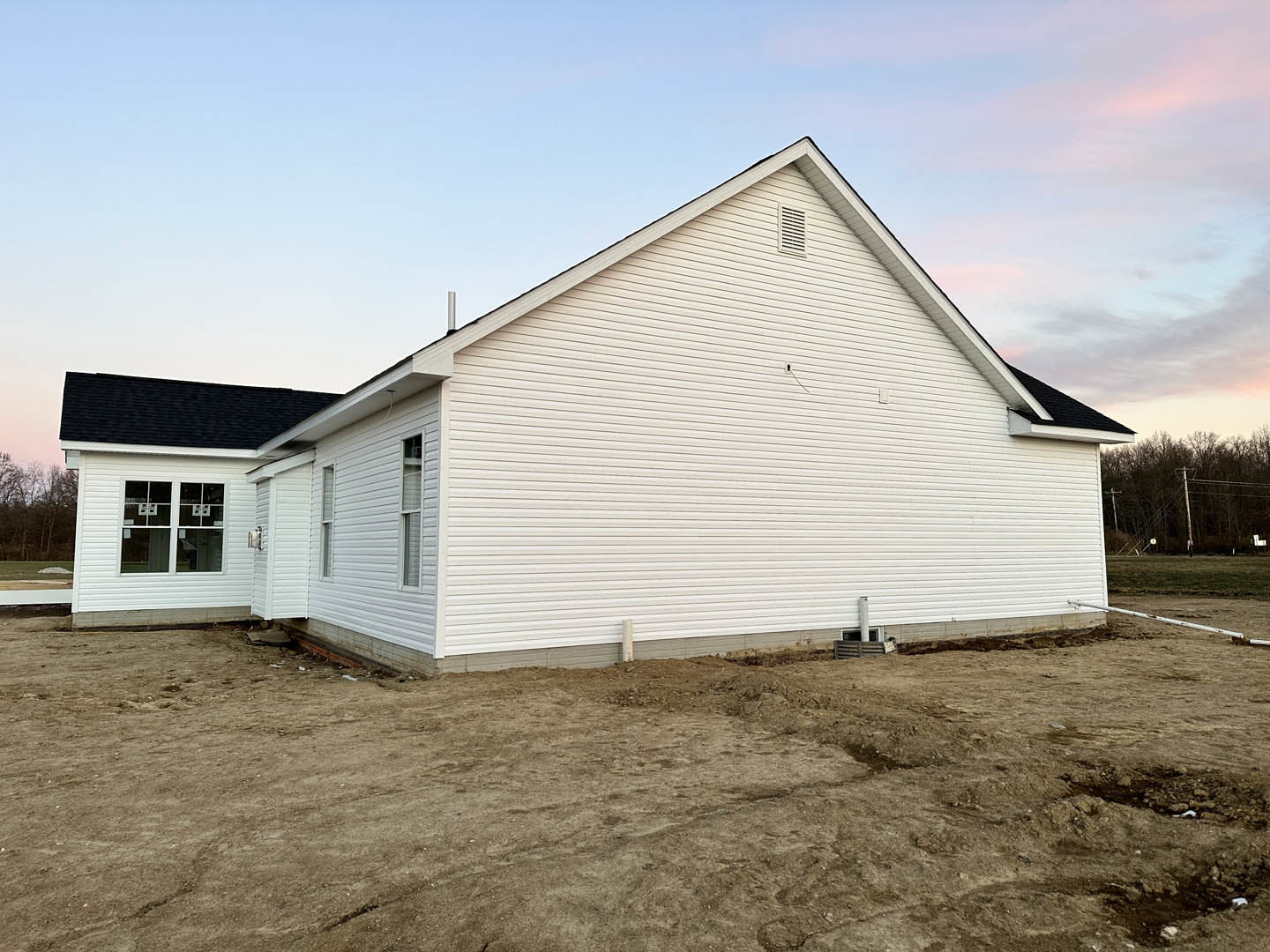 White house with black roof and chimney, dirt field in foreground, white siding, window displaying multiple signs, power line pole and wooded area in background, partly cloudy sky
