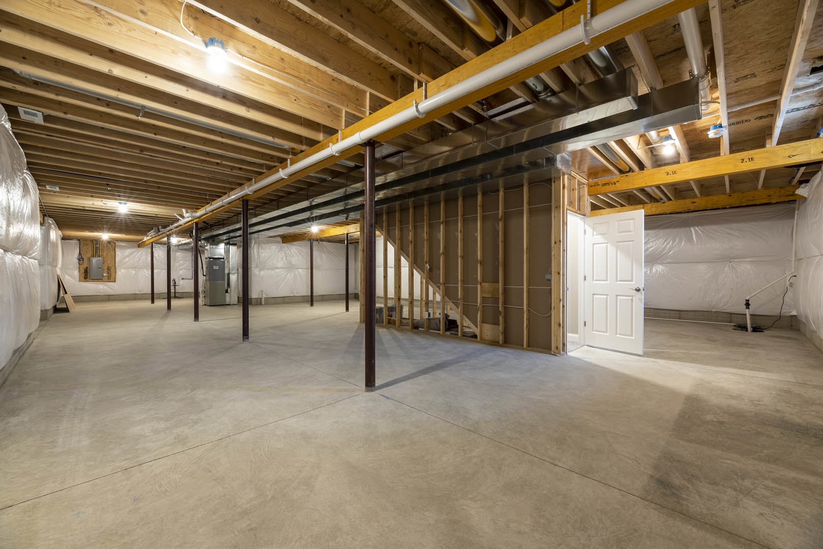 Concrete floor with central steel beam, wood ceiling featuring exposed pipe, white wall partially covered in plastic, white door with silver handle, gray refrigerator against wall