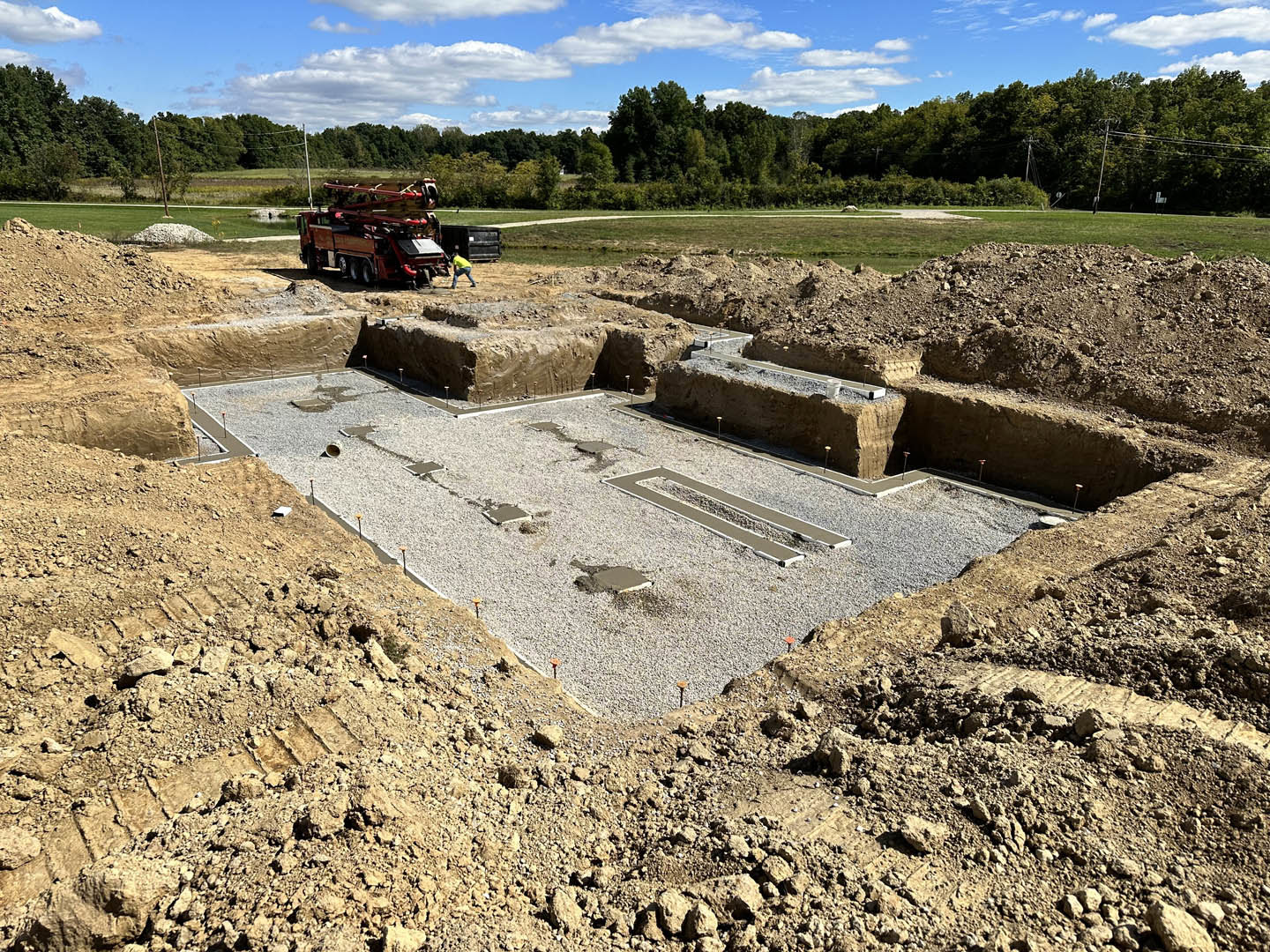Construction site with two trucks parked on dirt, red truck carrying pipes, muddy ground, scattered rocks, grassy patches, trees in background, blue sky with clouds overhead