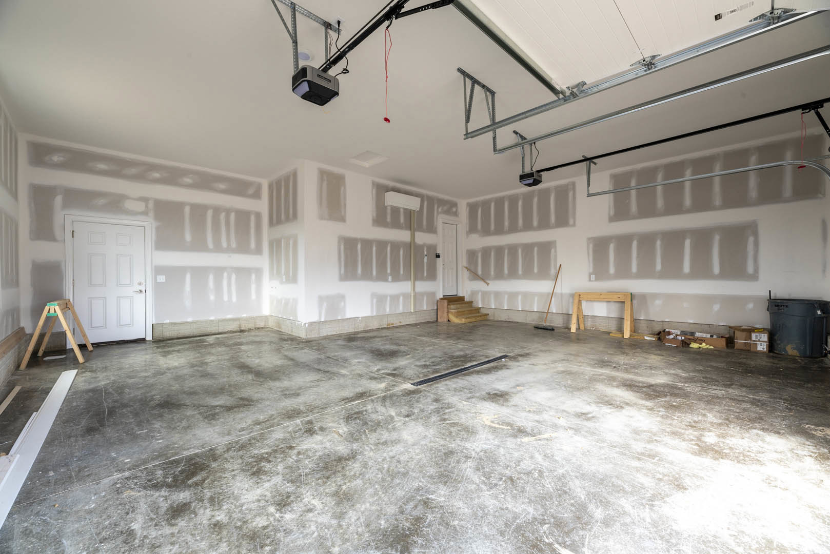 Concrete-floored room with white door, wooden ladder, floor drain, large black trash can, wood sawhorse, and exposed wood framing under plaster ceiling