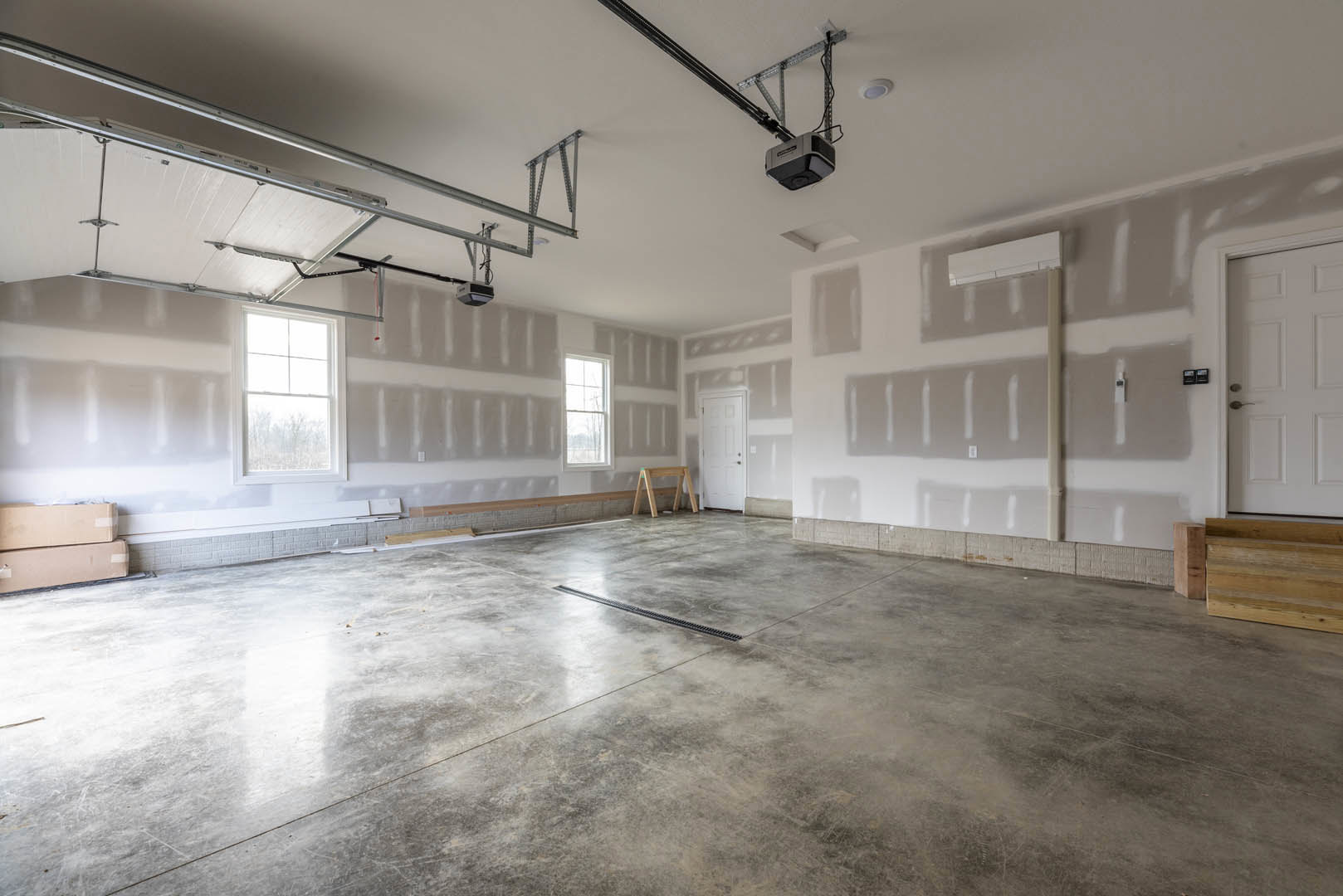 Concrete-floored room with a floor drain, white-framed window, white door with handle, wooden sawhorse, and wooden box near the door.