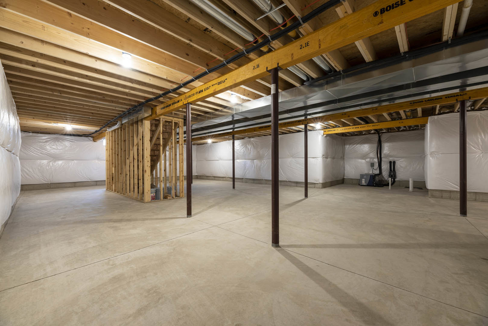 Exposed wooden ceiling beams, concrete floor with metal support poles, large white plastic-wrapped bag, grey box with black and blue cover, unfinished walls in an empty basement