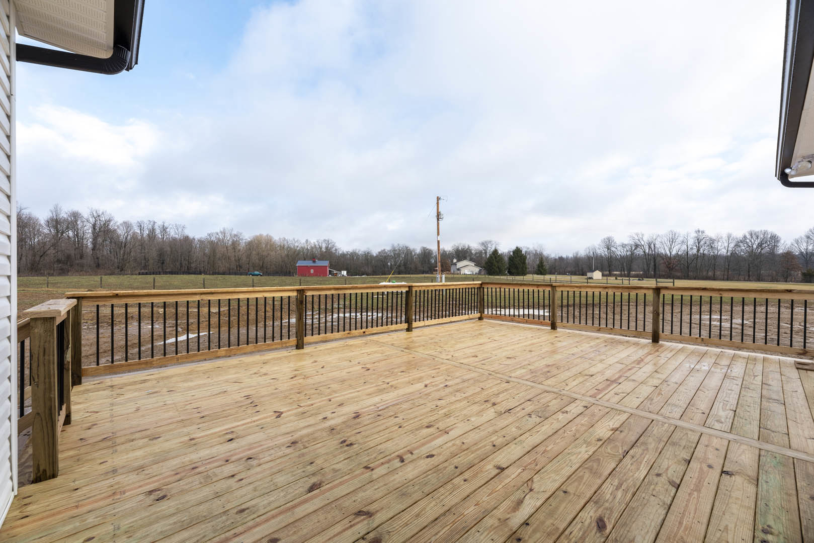 Wooden deck with black metal railing, fenced perimeter, and leafy trees in the background under a partly cloudy sky