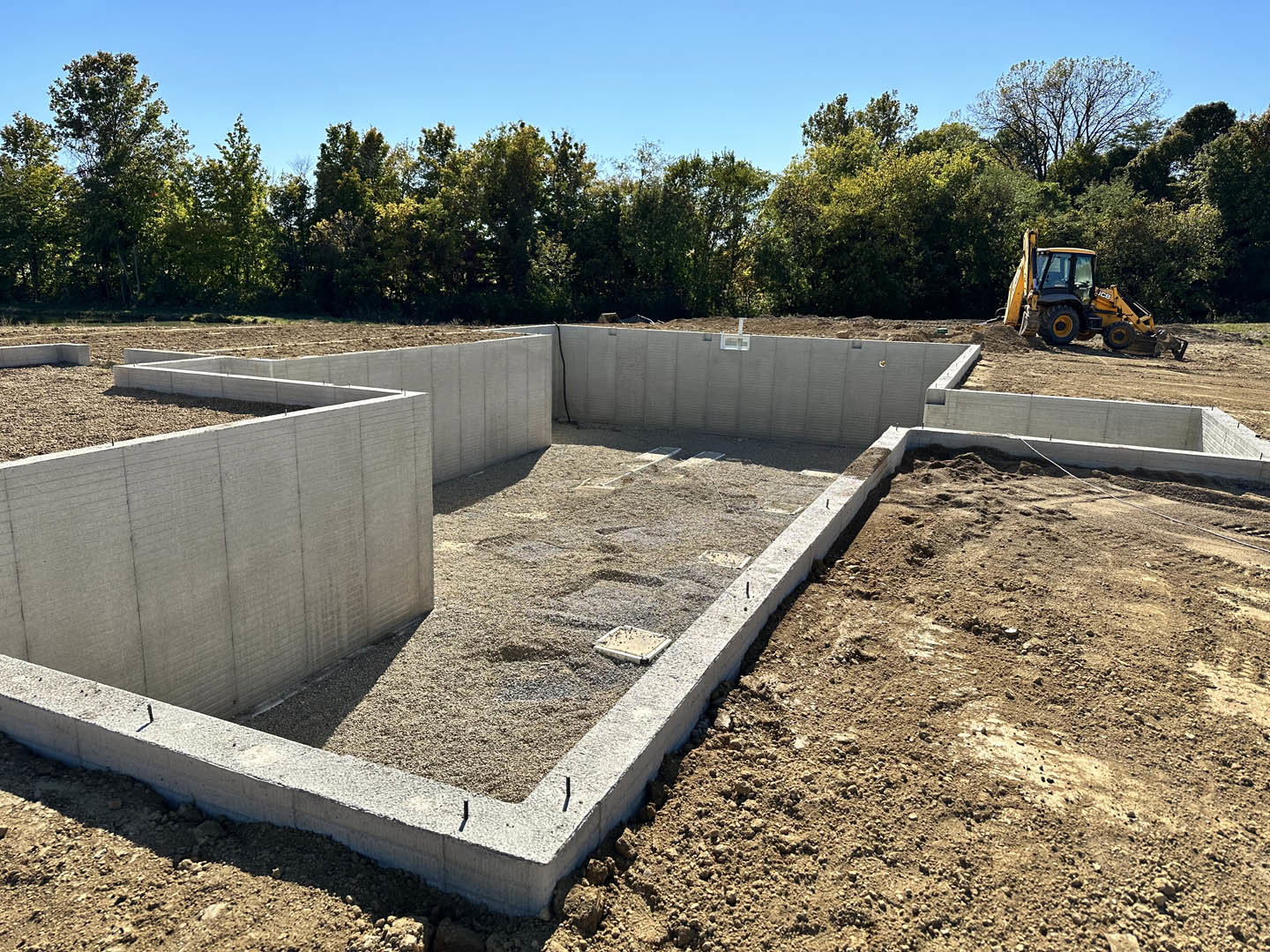 Bulldozer parked beside freshly poured concrete foundation surrounded by gravel, dirt, and construction materials; trees and blue sky in background