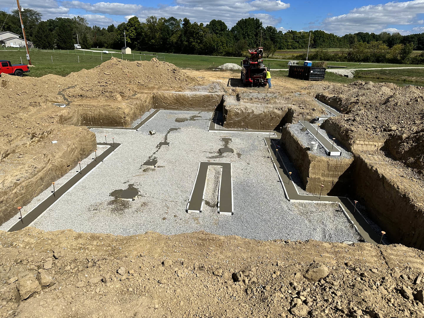 Dirt construction site with a yellow tractor, red truck, and scattered rocks; trees and cloudy sky in the background
