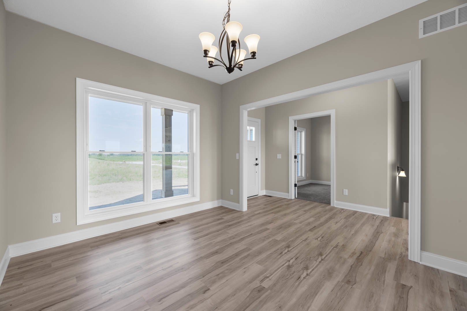Wood flooring in a spacious room with white walls, decorative molding, a five-light chandelier, large window overlooking a field, and a floor vent near the baseboard.