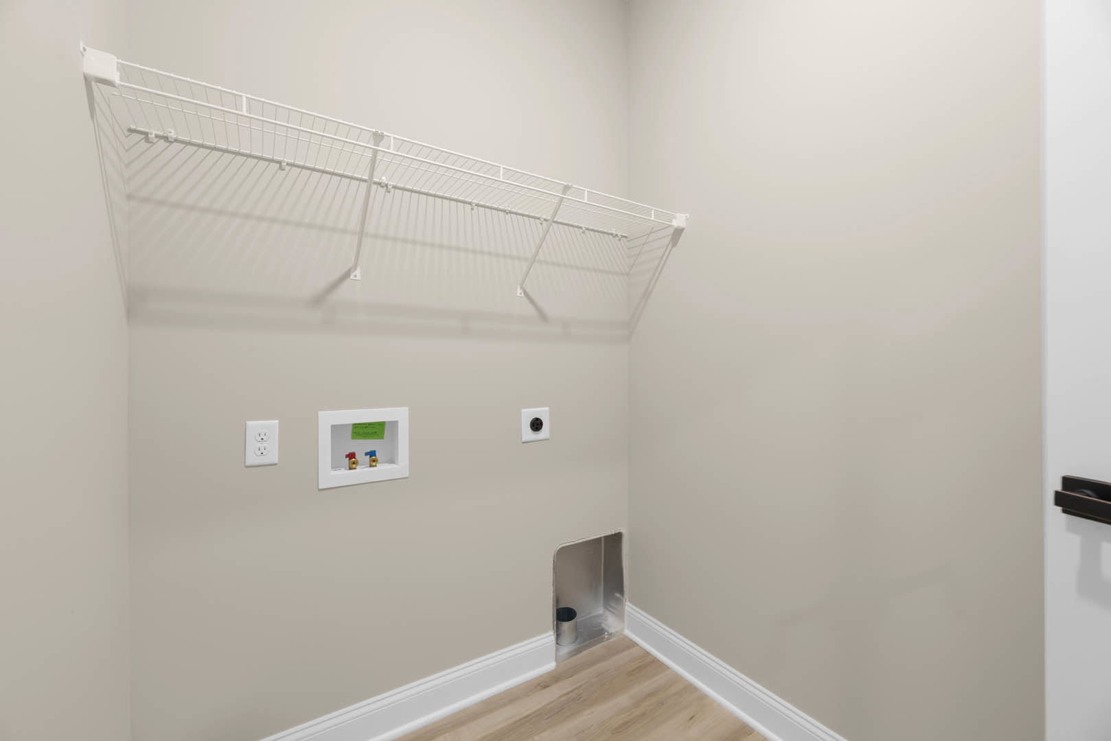 White wire rack mounted on plaster wall above white electrical outlet, metal canister nearby, green sign on framed white box, white shelf against white background.