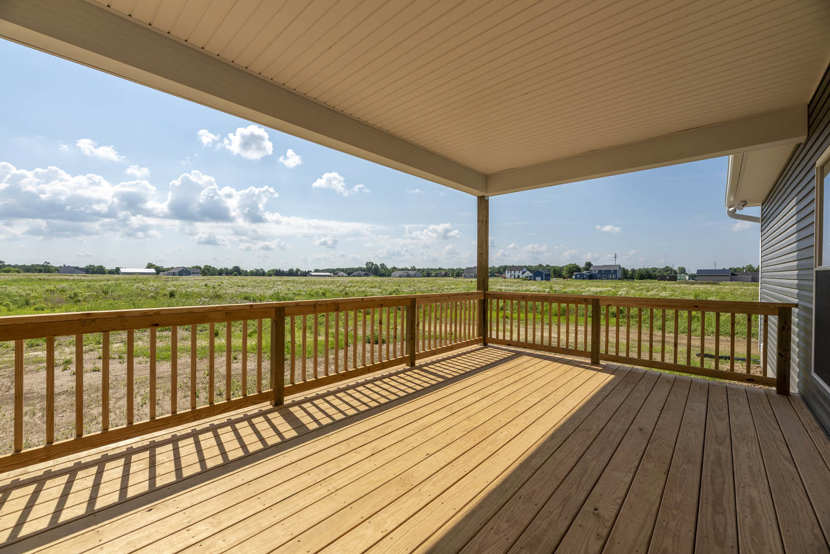 Wooden deck with horizontal railing, expansive grassy field beyond, blue sky with scattered clouds overhead
