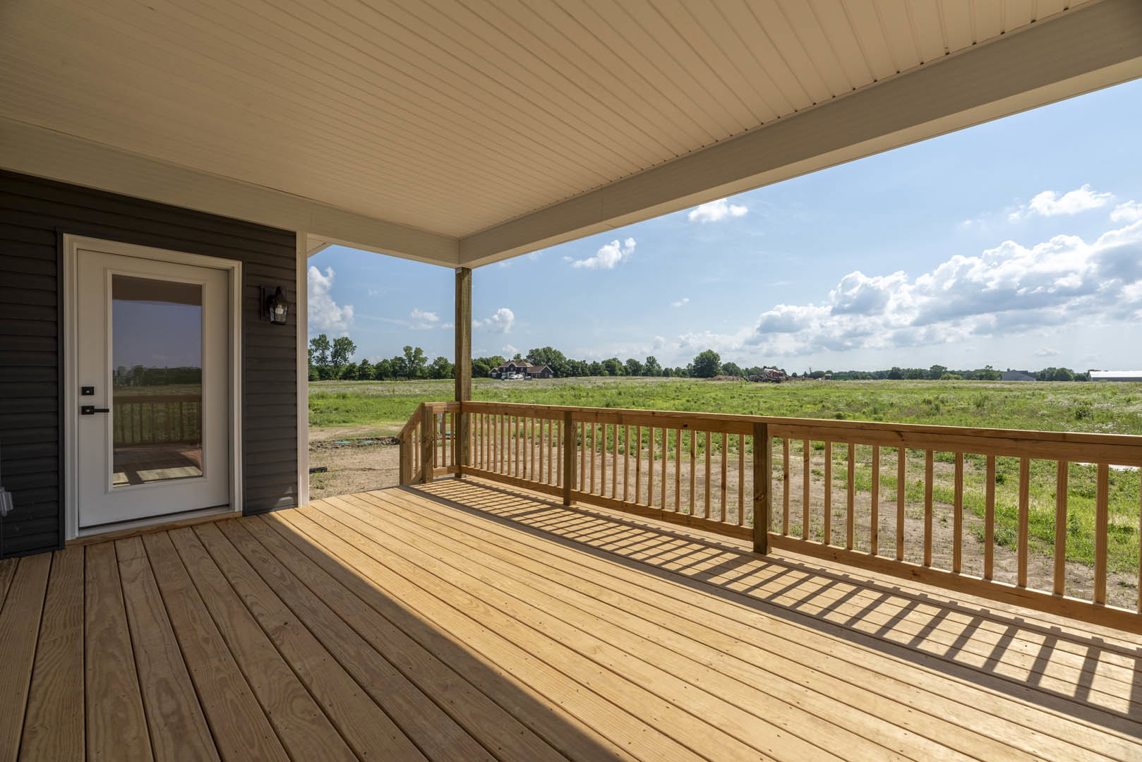Wooden deck with railing overlooking grassy field, white roof overhead, trees and sky in background