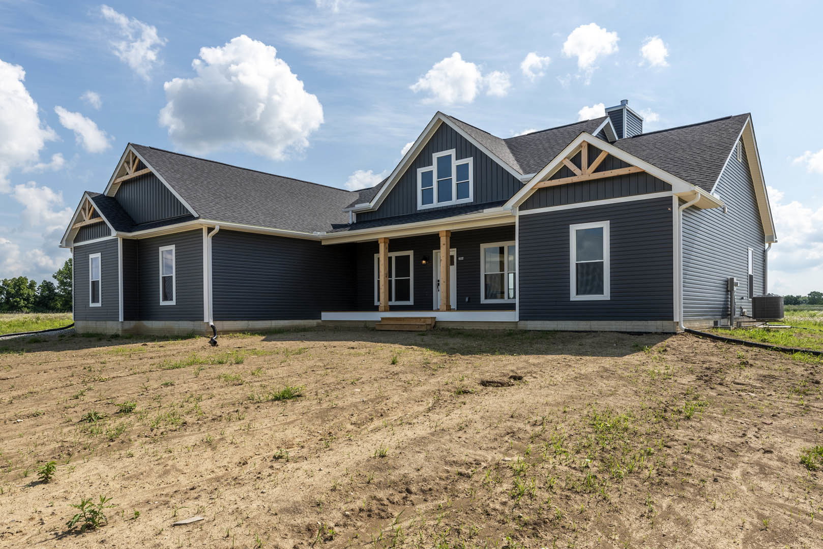 Two-story house with white-framed windows and covered porch, surrounded by a dirt field with patches of grass, under a partly cloudy sky; large black box visible on a truck in