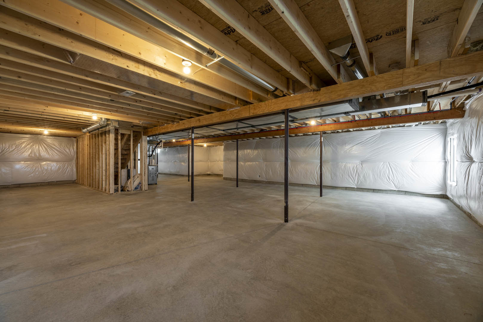 Spacious room with exposed wood ceiling beams, metal support poles, concrete floor, white wall partially covered with plastic sheeting, and single light bulb hanging from ceiling