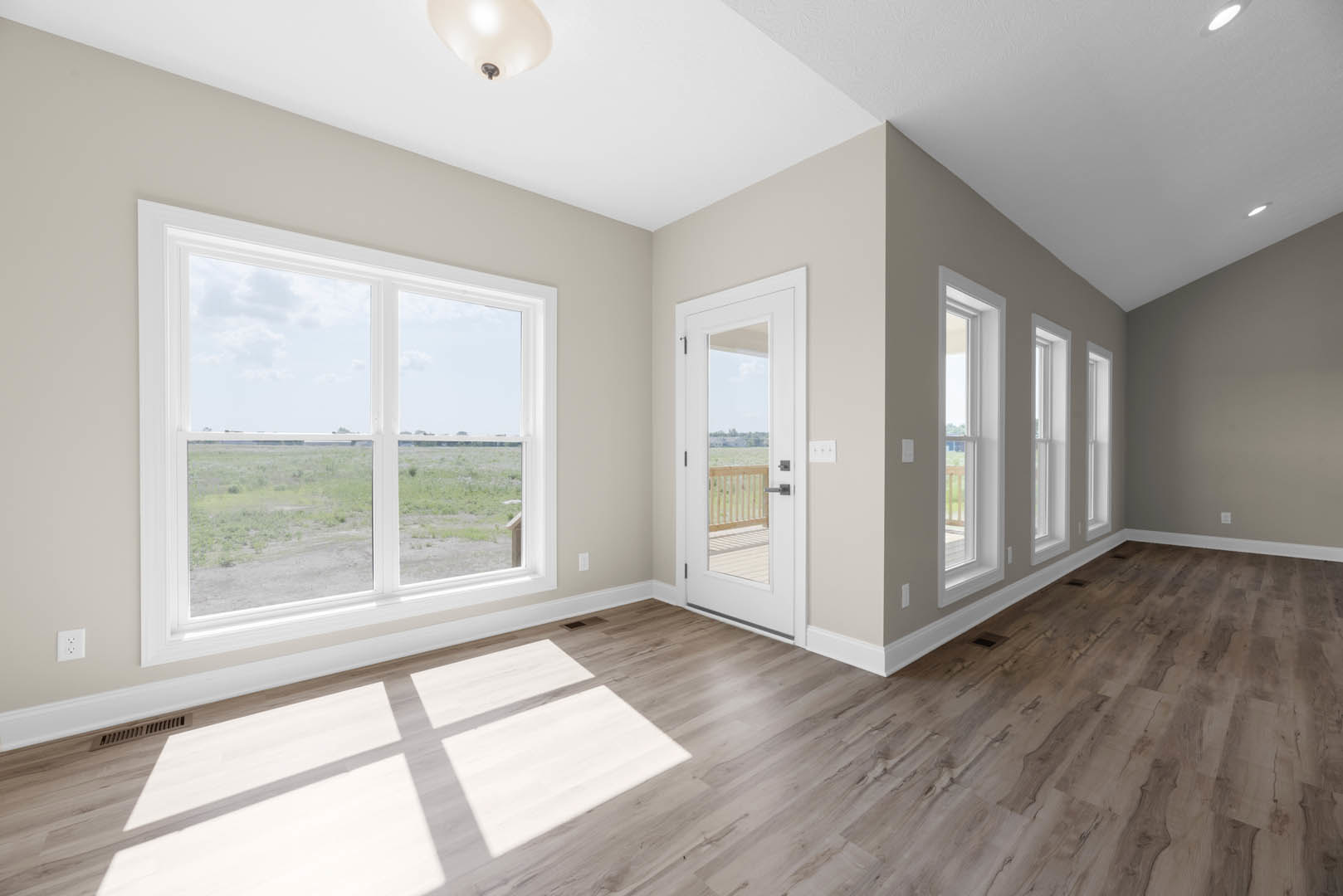 Bright room with wood flooring, white walls, large windows, and a white door overlooking a grassy field; modern light fixture hangs from ceiling.