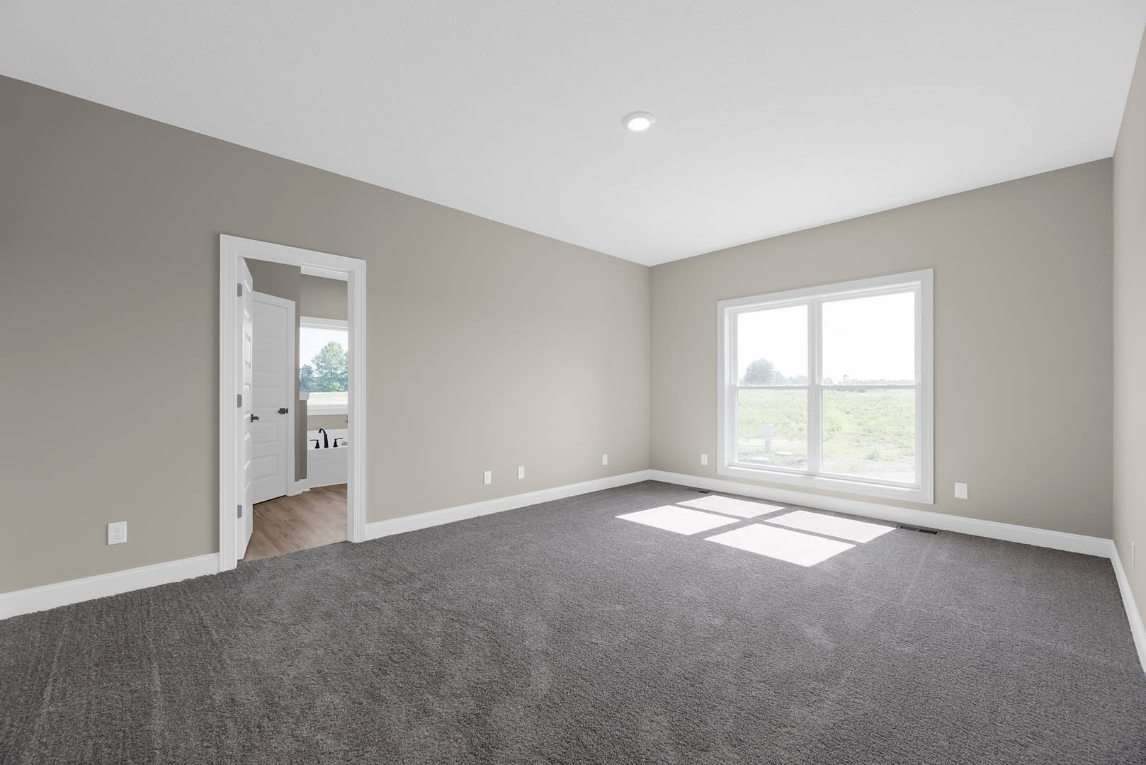 Grey carpeted room with white walls, large window overlooking a grassy field, white door with black hardware, simple ceiling and baseboard molding.