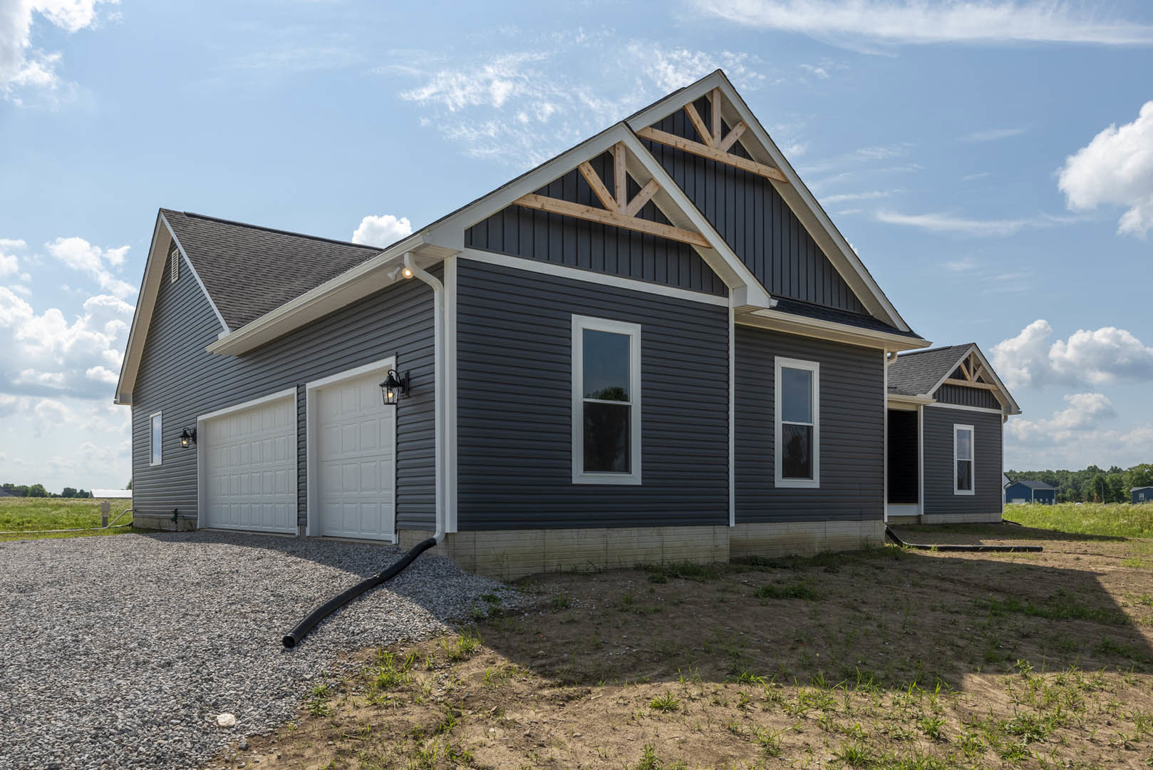 Modern home exterior with white garage doors, gravel driveway, black garden hose, white-framed windows, gray roof, and partly cloudy sky.