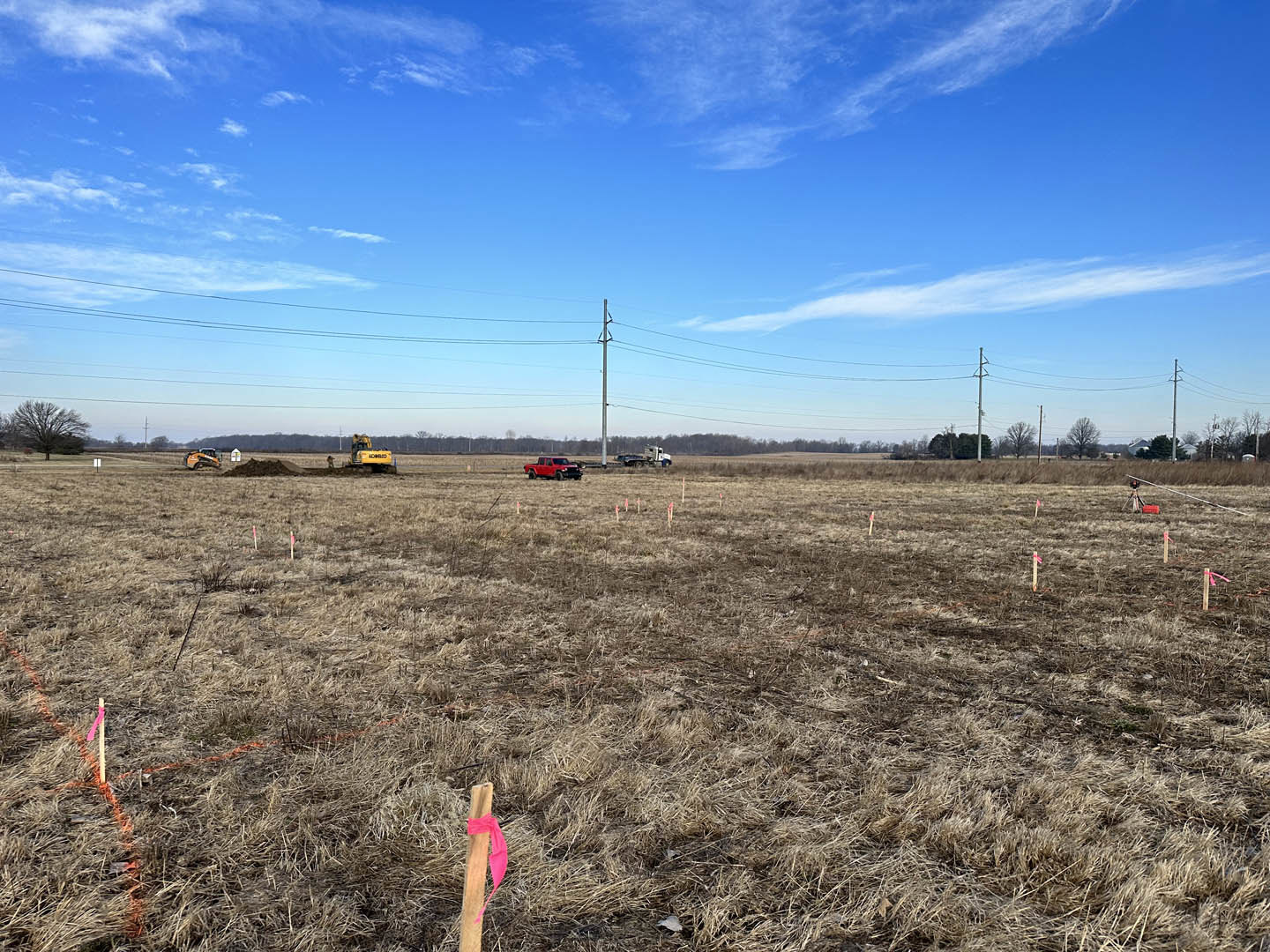 Grassy field with scattered vehicles, yellow bulldozer, wooden post with pink ribbon, power lines overhead, tree in background, blue sky with clouds