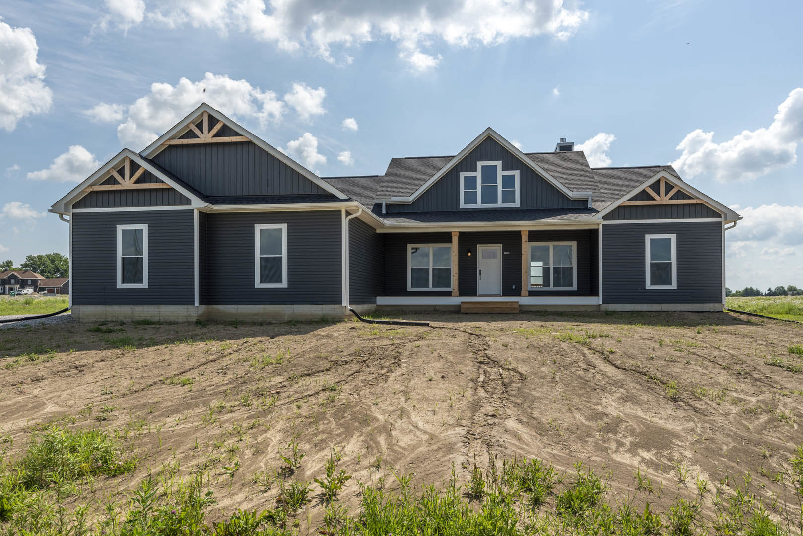 White single-story house with gray roof, covered porch, white framed windows, and white front door with window, set beside a dirt field with patches of grass and a visible pipe