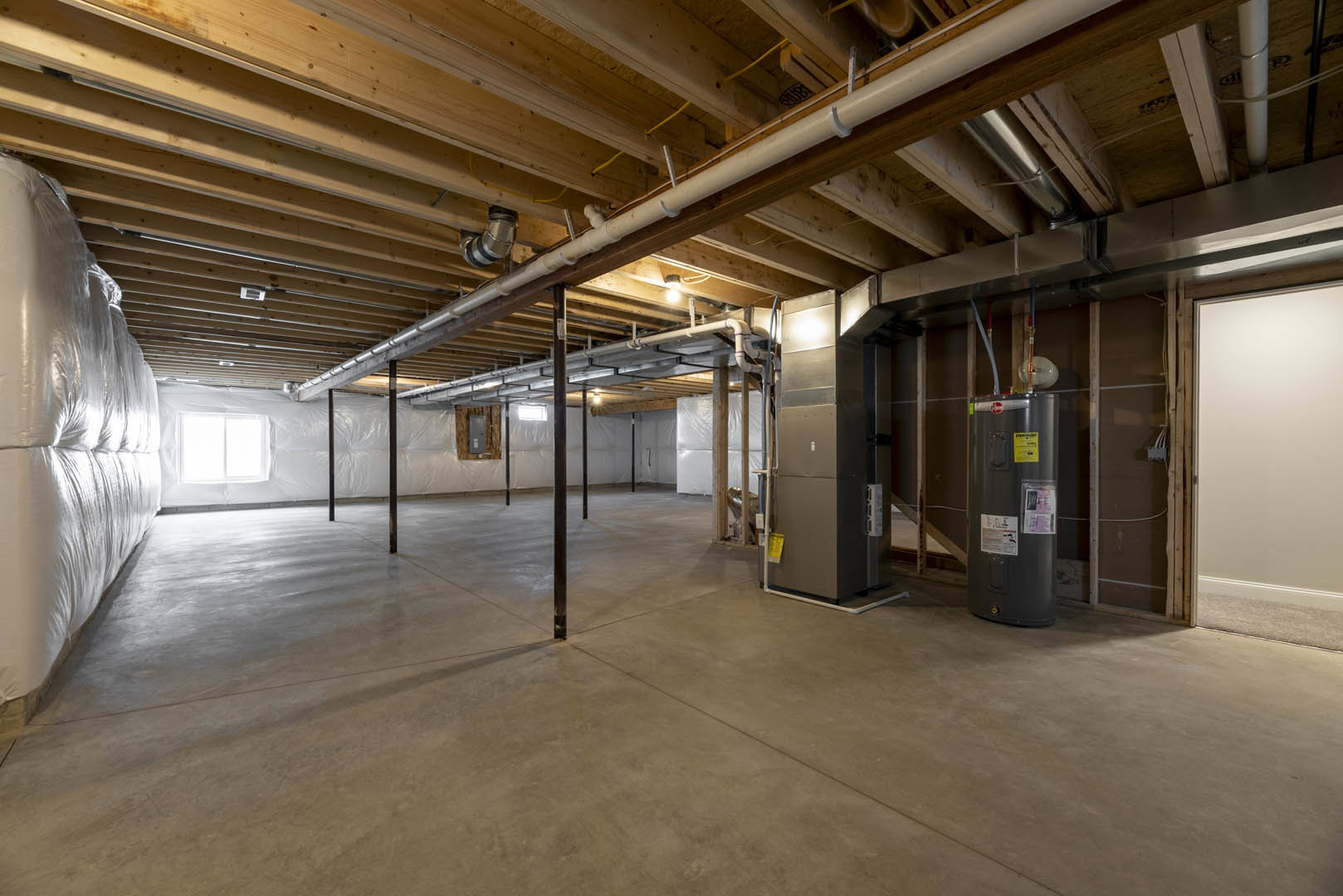 Basement room with exposed steel and aluminum pipes along the ceiling, concrete floor, white wall with mounted light fixture, black cylinder, metal pole, and white cushion.