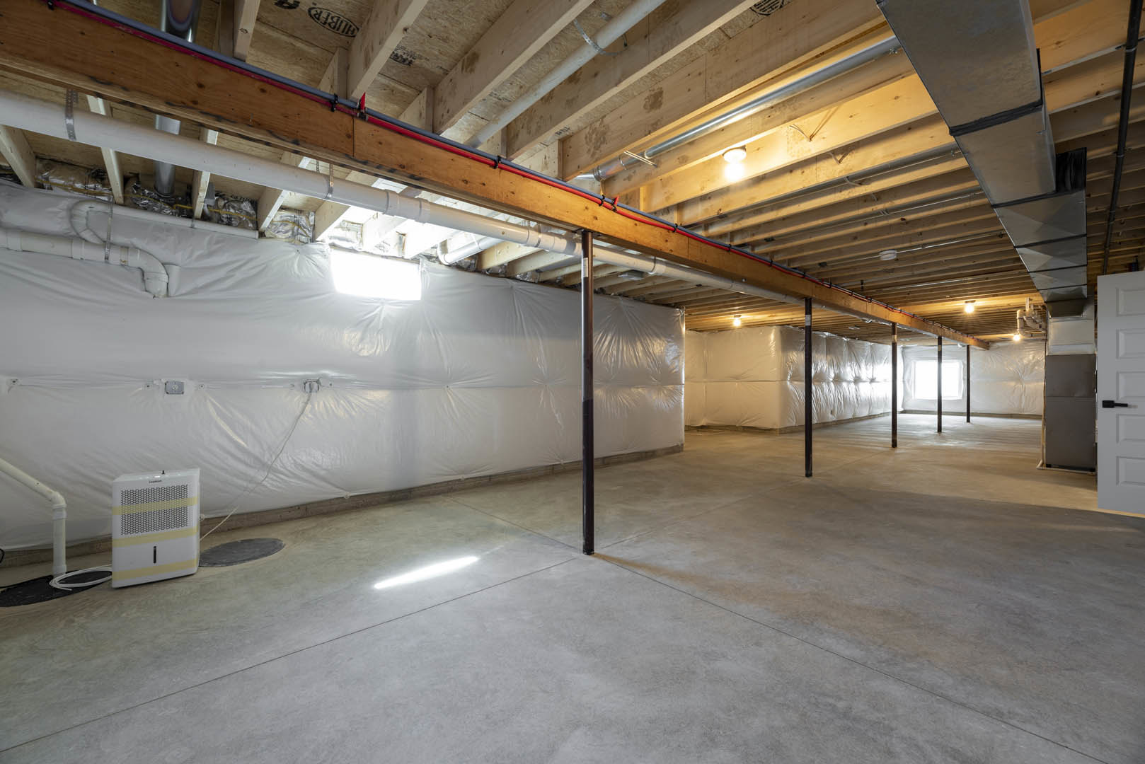 Basement room with white plastic coverings draped over walls, exposed wooden beam with pipes, concrete floor with a metal pole, and a white square device featuring yellow stripes.