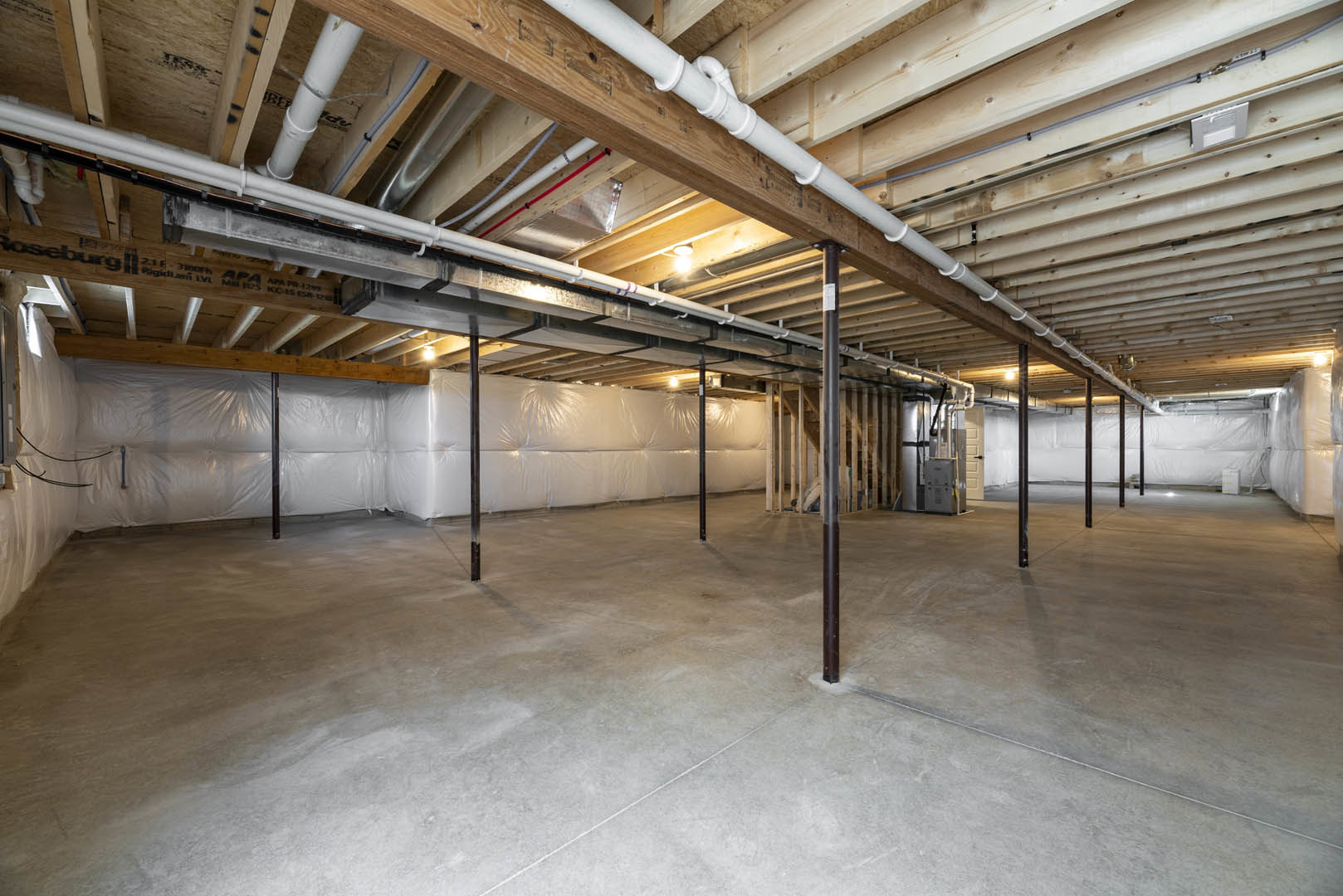 Exposed wooden ceiling with white pipes, concrete floor supported by metal poles, white rectangular object with handle, and stacked white bags in unfinished basement.