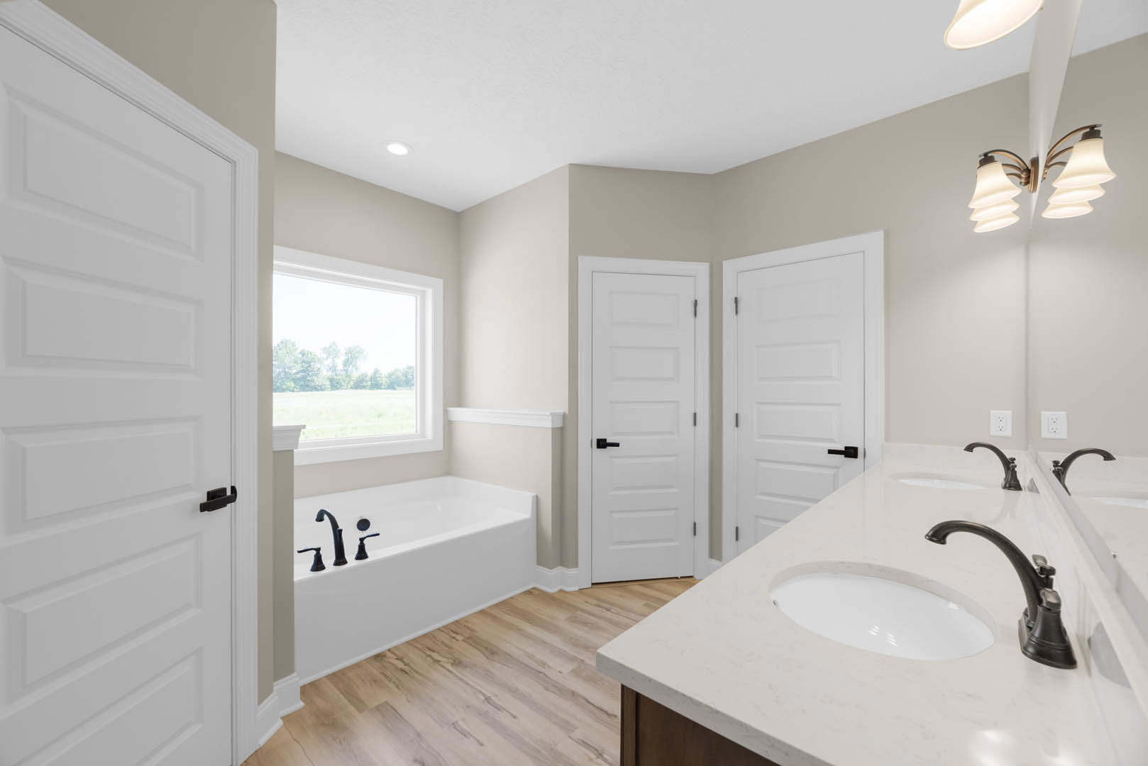 Bathroom with double sinks, white cabinetry, freestanding bathtub, large window overlooking trees, black hardware on white door, modern light fixture, and neutral tile walls.